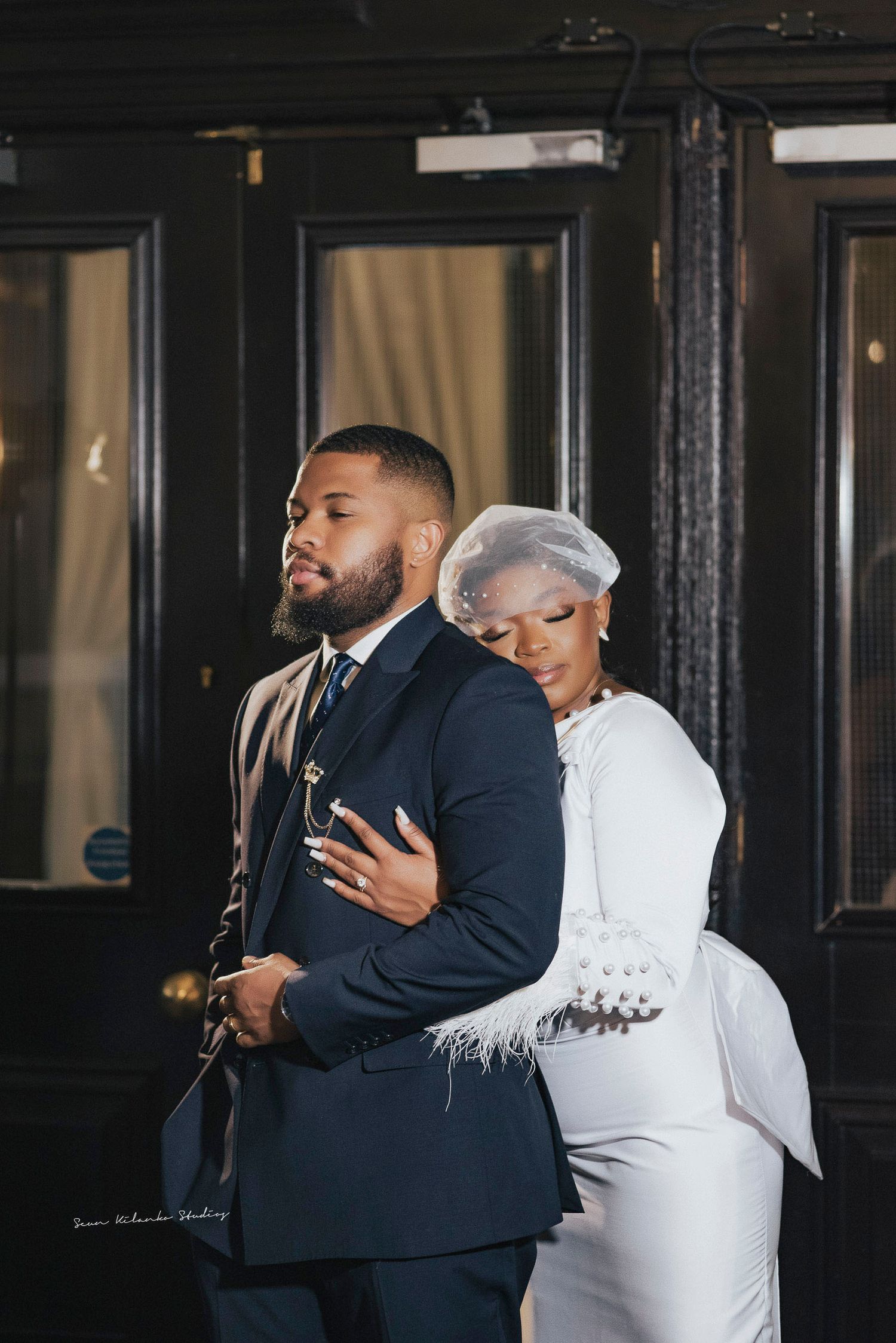 Stylish couple in navy suit and white feathered dress pose against black doorway.
