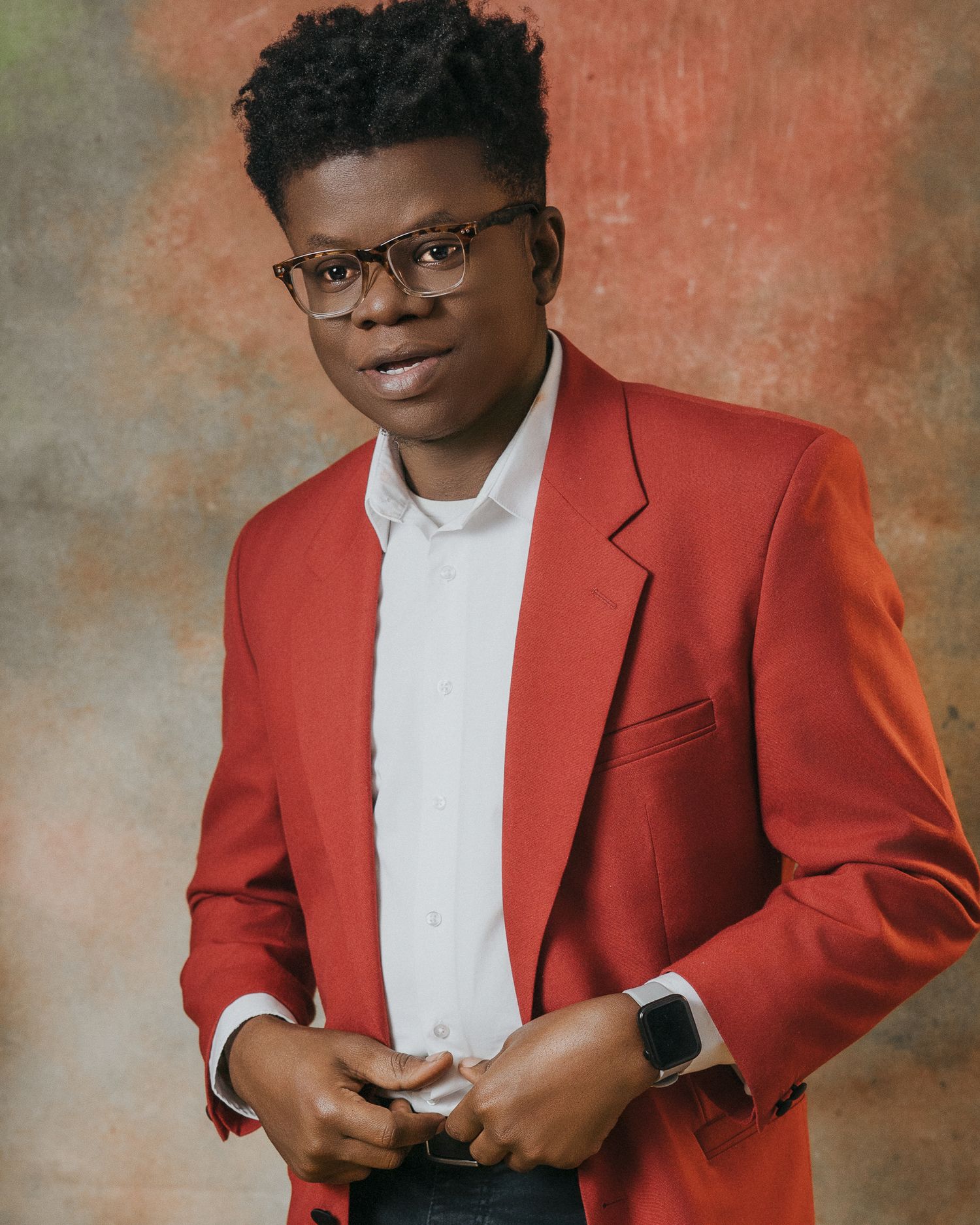 A person in a bright red blazer and glasses poses against a textured brown backdrop in a professional portrait series.