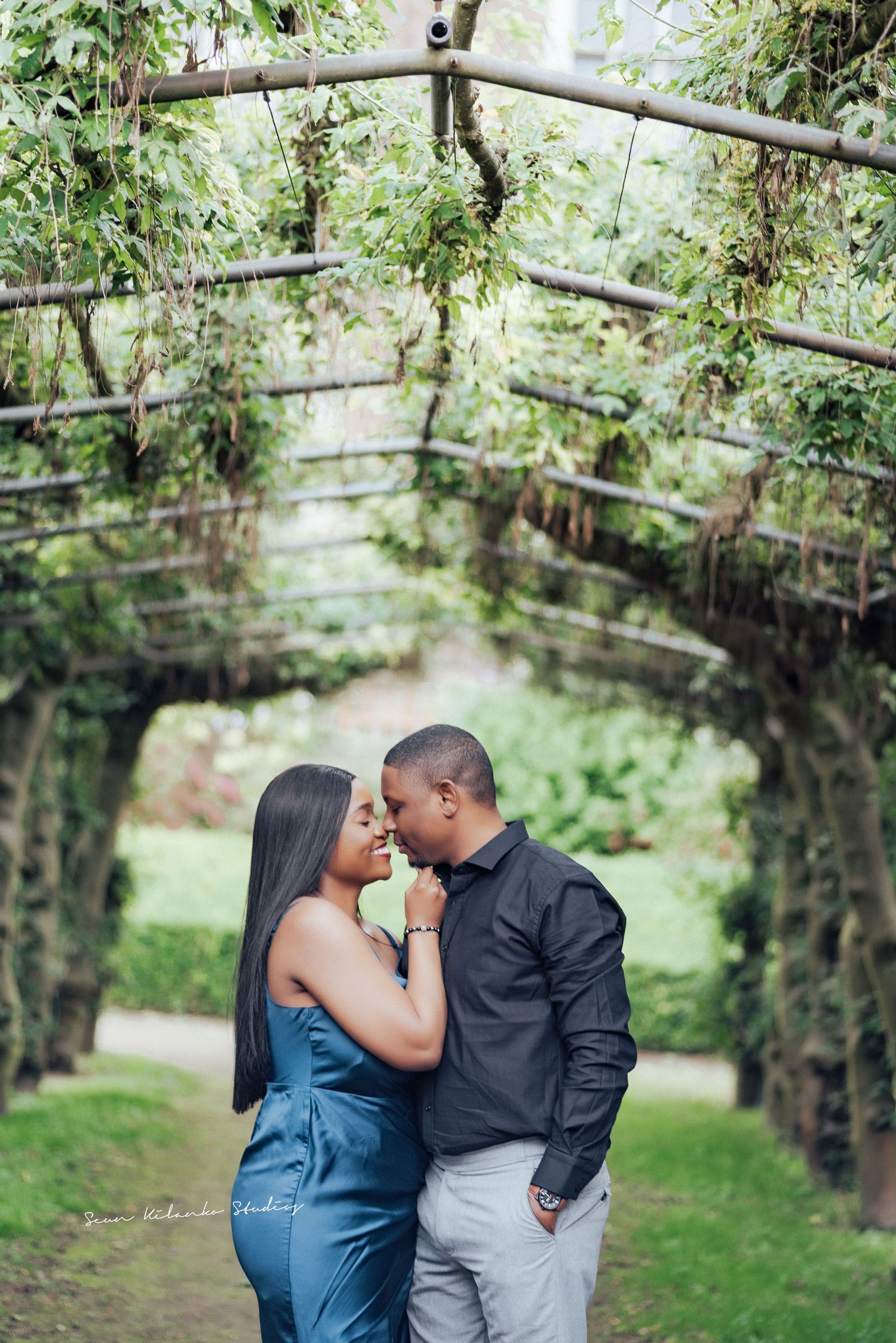A romantic couple embraces under a garden archway covered in green vines and foliage.