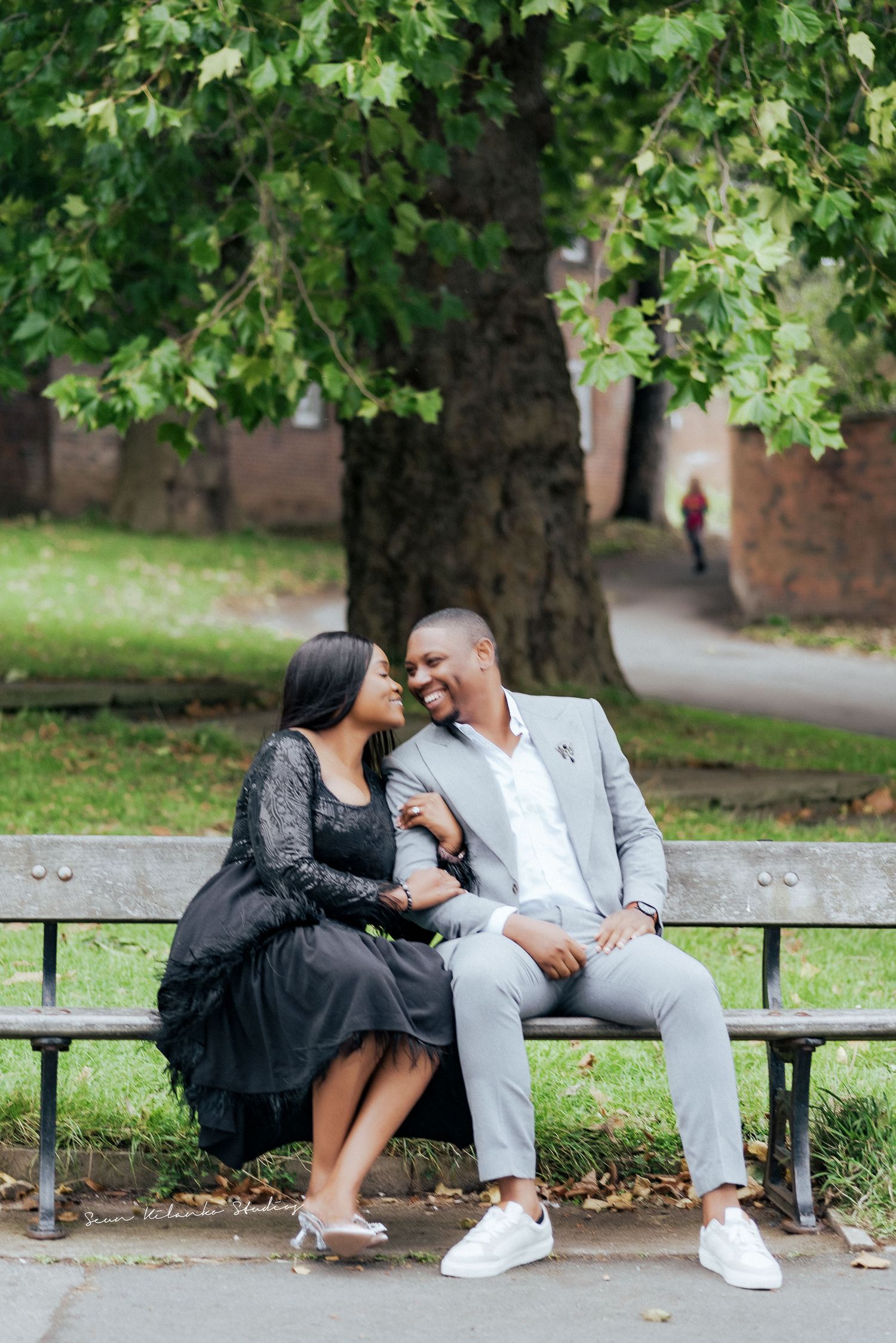 Couple shares tender moment while sitting on park bench under green tree.