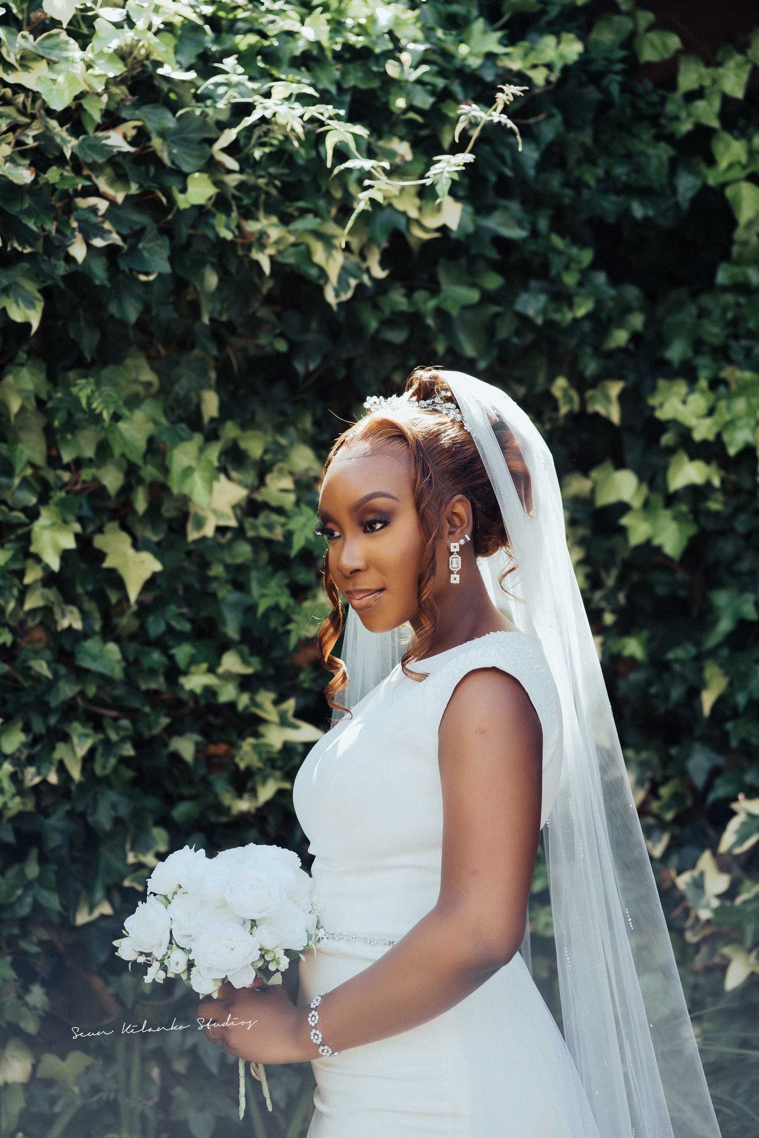 Bride with white bouquet poses in sleek dress against green foliage.