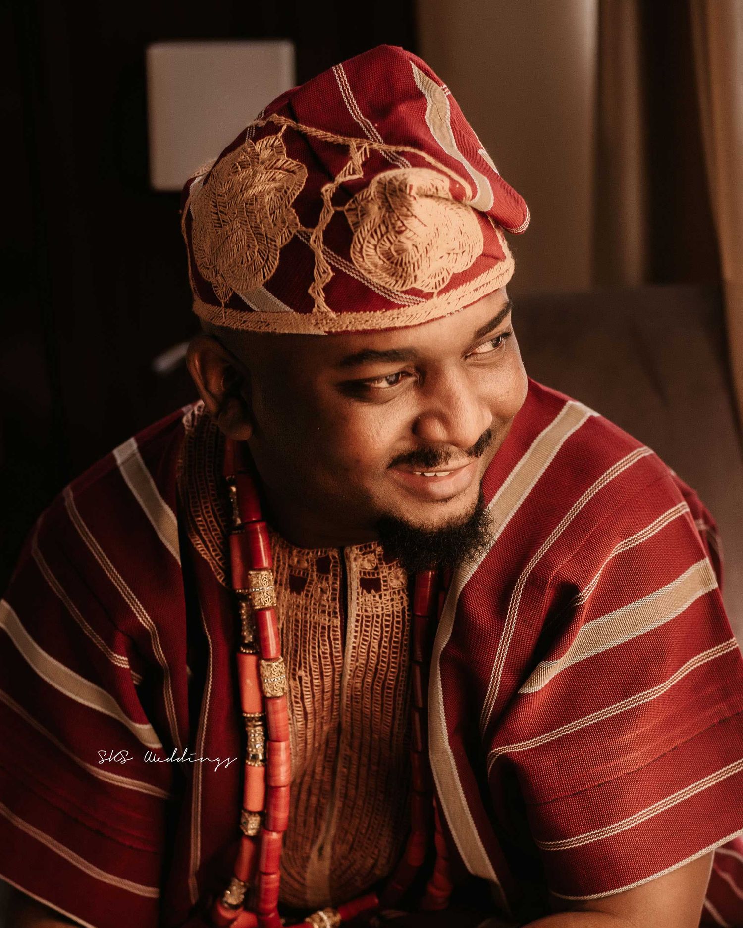 Person wearing traditional Nigerian Yoruba attire including a red and gold cap and striped agbada in warm lighting.