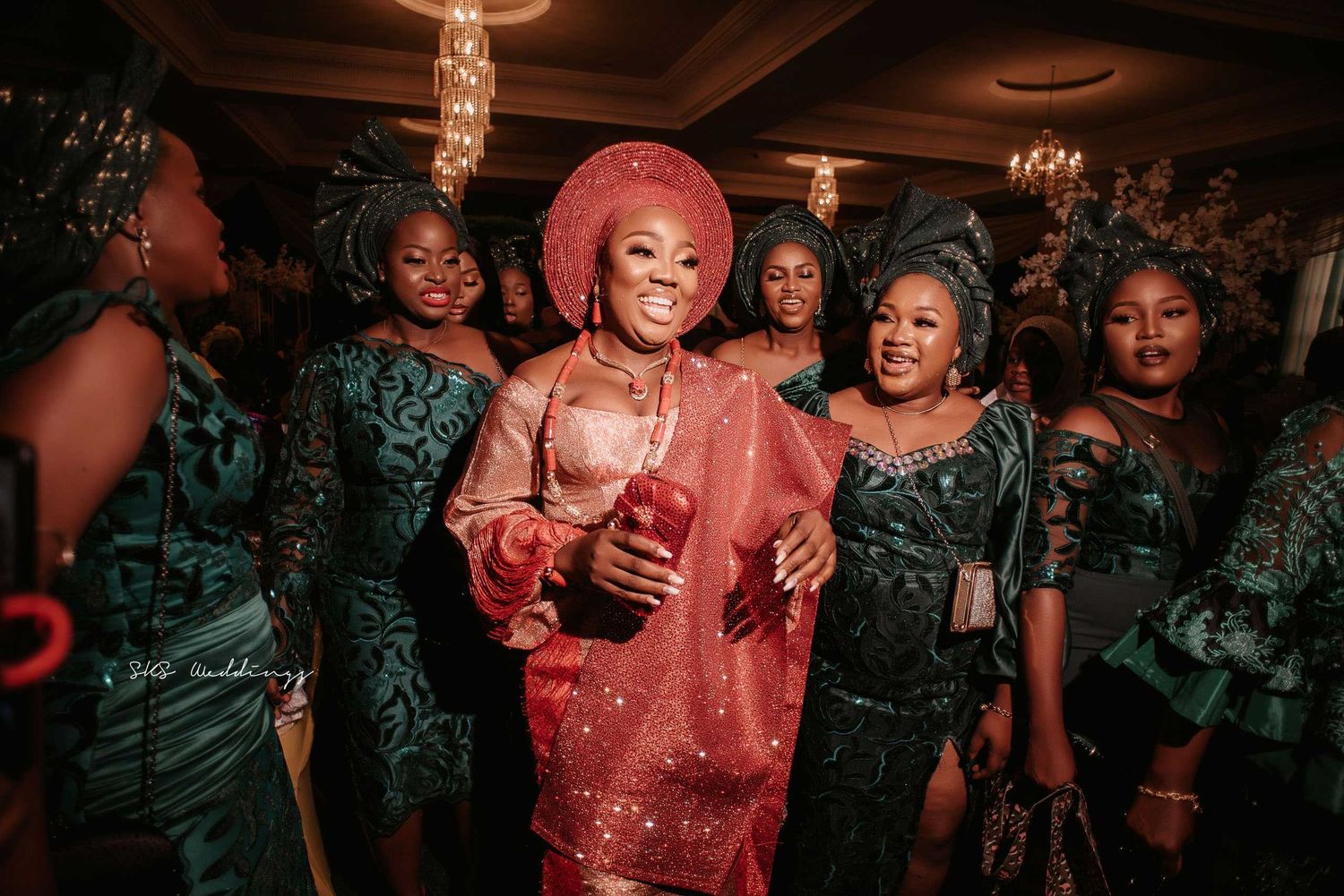 A celebratory group at a wedding reception wearing emerald green and coral pink attire under warm lighting.