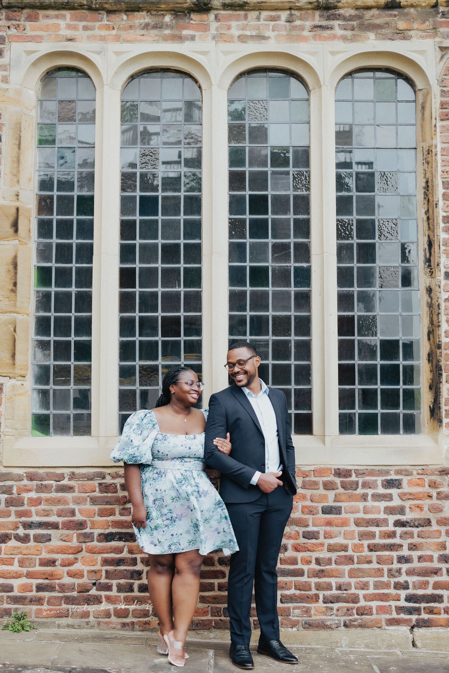 Stylish couple poses against historic church windows and brick wall.