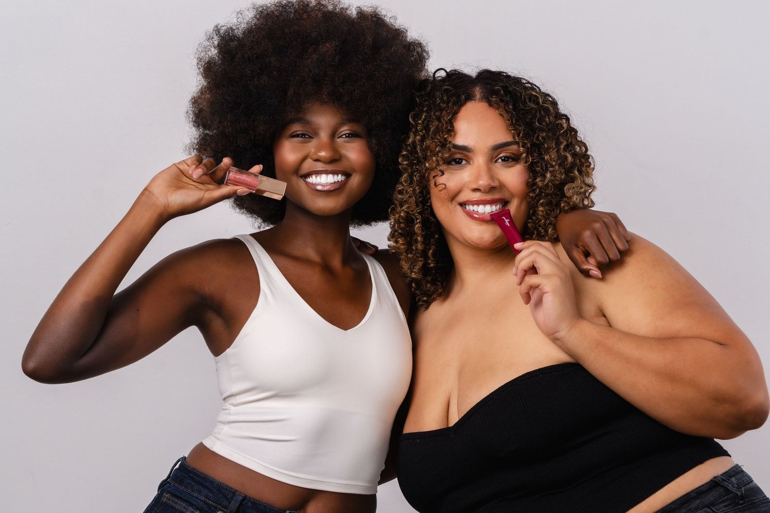 Two smiling women posing together, one in a white crop top and one in a black top, against a gray background.