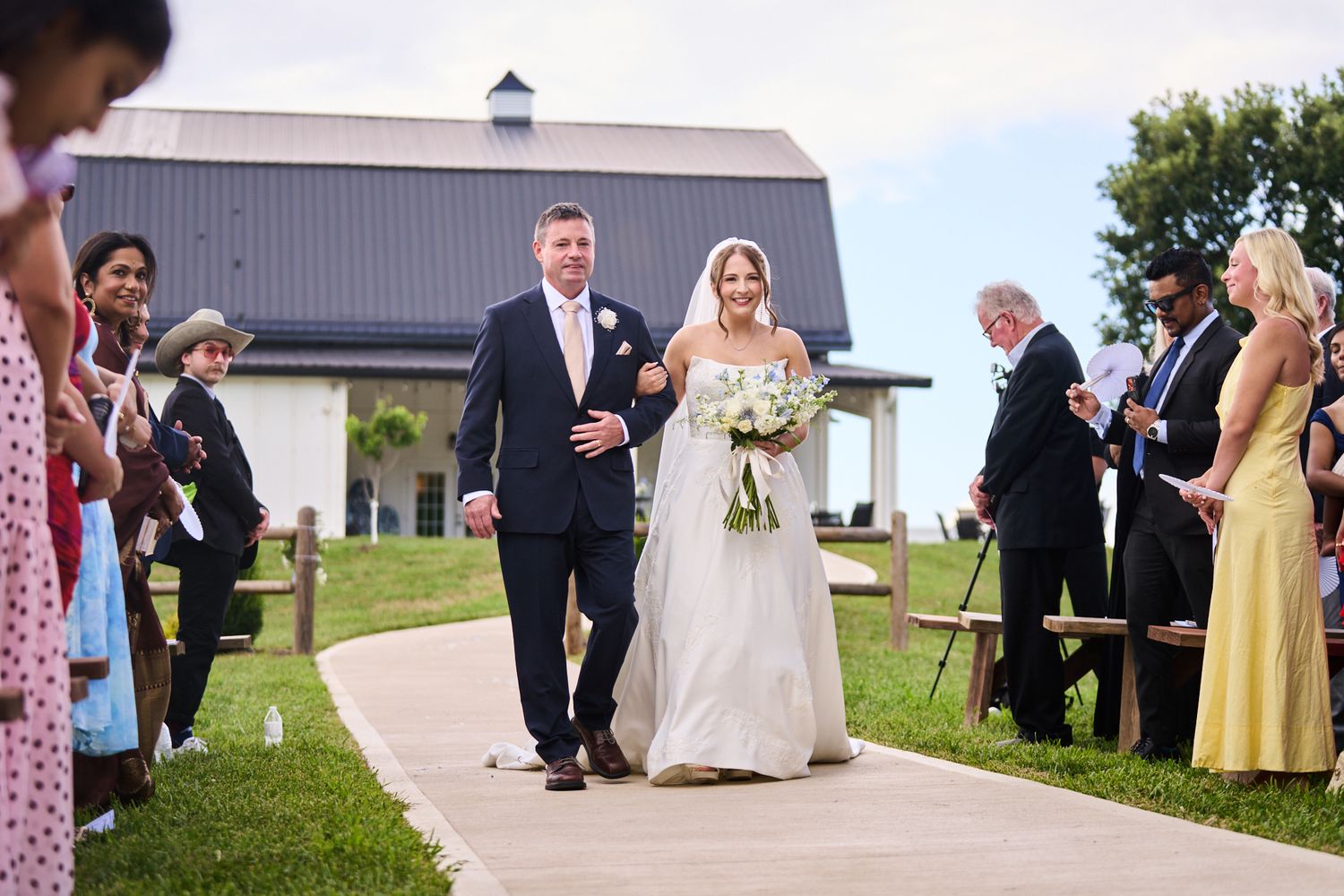 A bride in a white gown carrying a bouquet walks down the aisle on the arm of a man in a navy suit at an outdoor barn wedding ceremony.