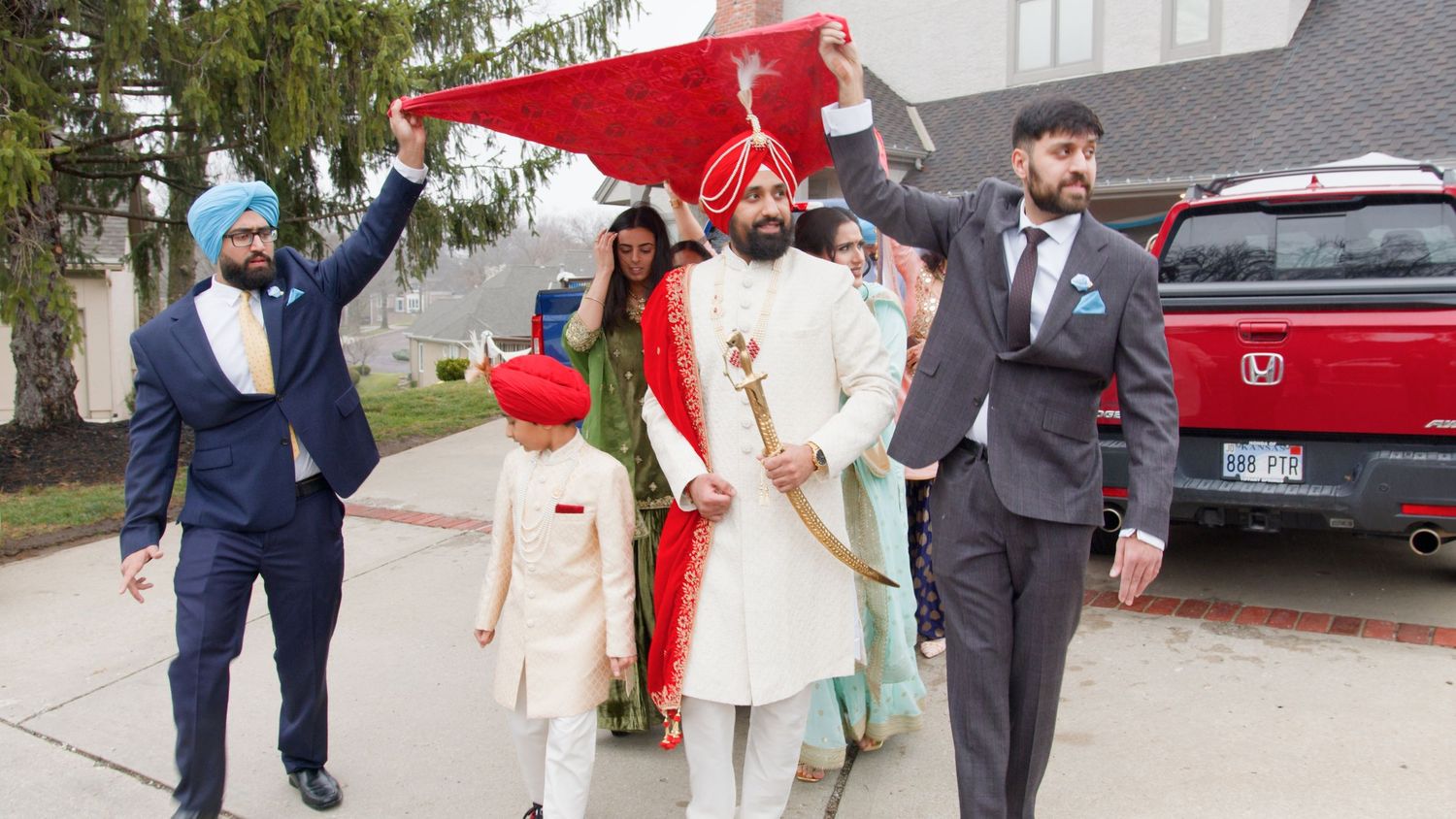 Groom in white sherwani and red turban holds a sword while men hold a red cloth canopy overhead during a Sikh wedding procession outdoors.