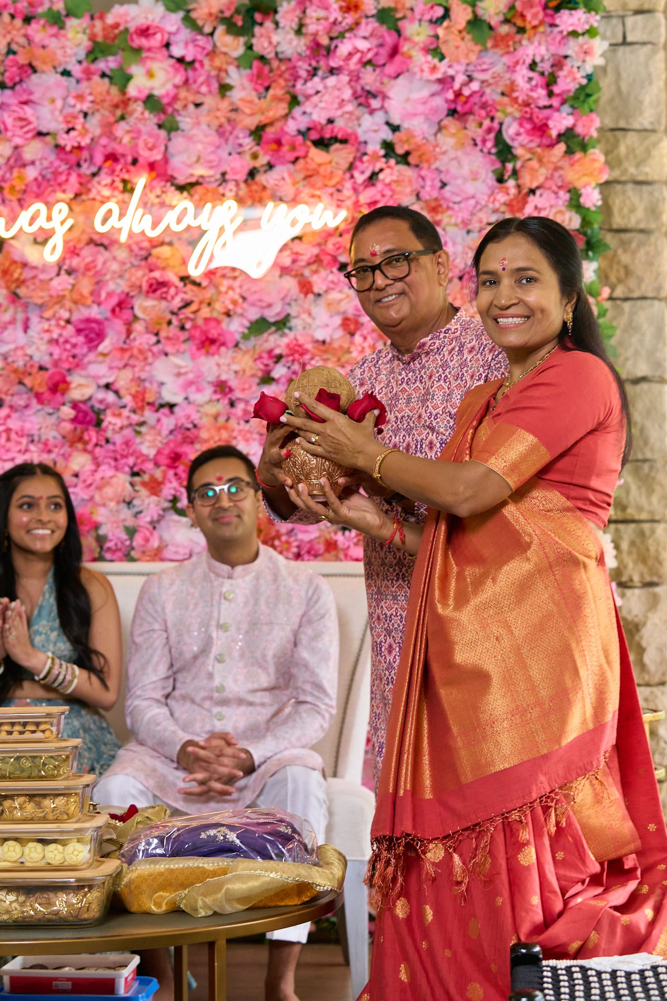 Woman in an orange silk saree and man in glasses hold a gift at an Indian ceremony with a pink floral wall and guests seated nearby.