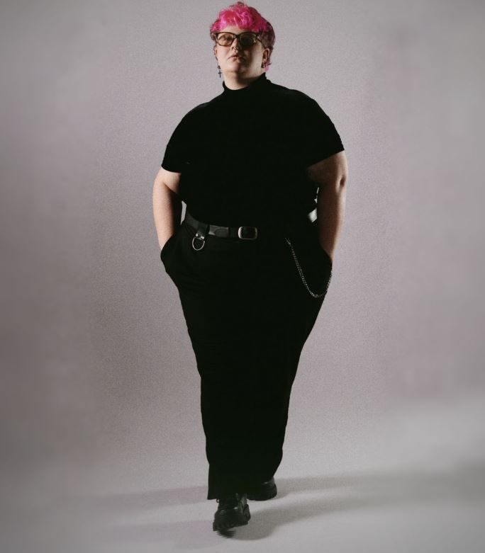 Person with short pink hair and glasses stands in a black turtleneck, trousers and boots with hands in pockets against a gray studio backdro
