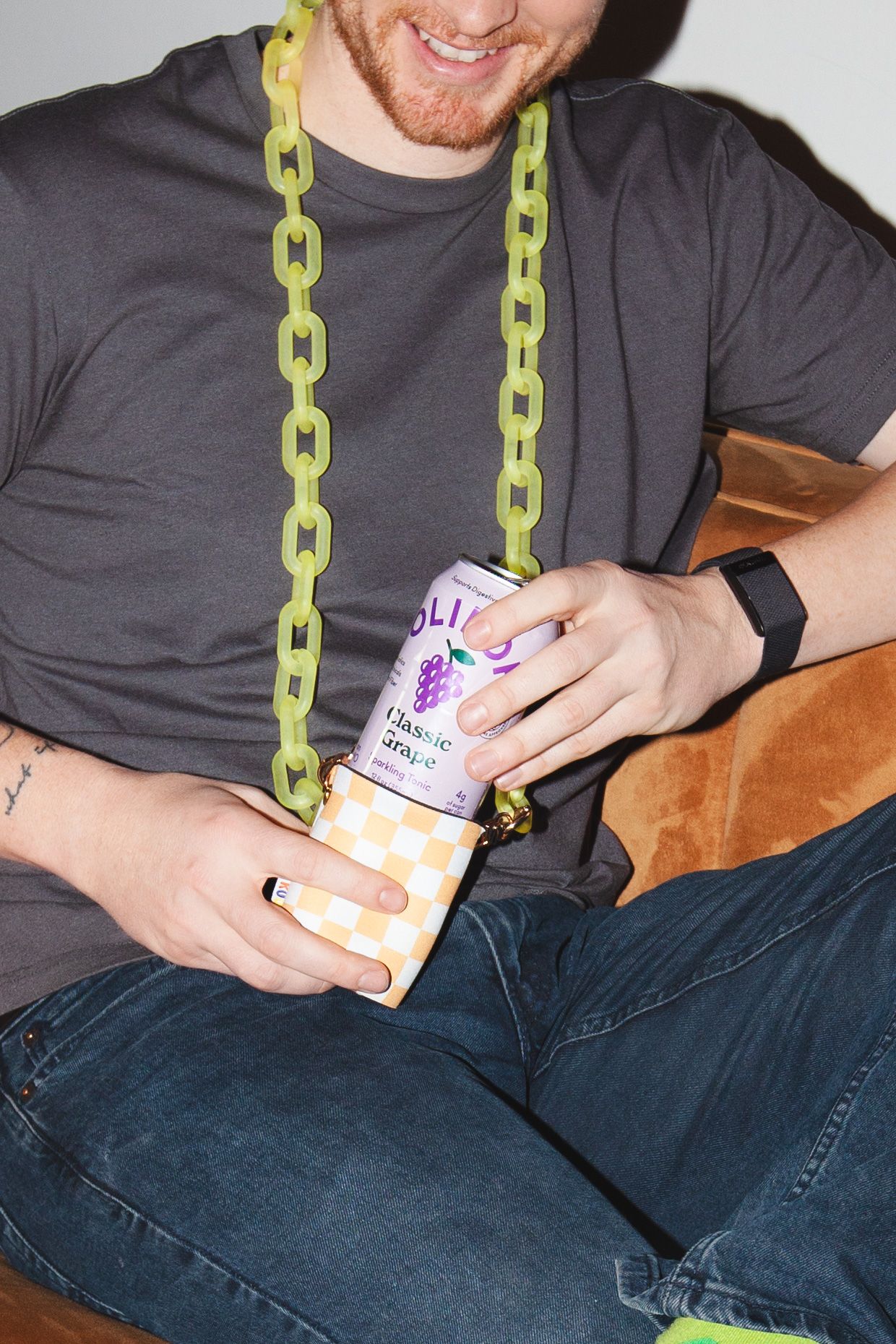Man wearing green chain necklace holding a grape-flavored Olipop soda can in a checkered koozie.