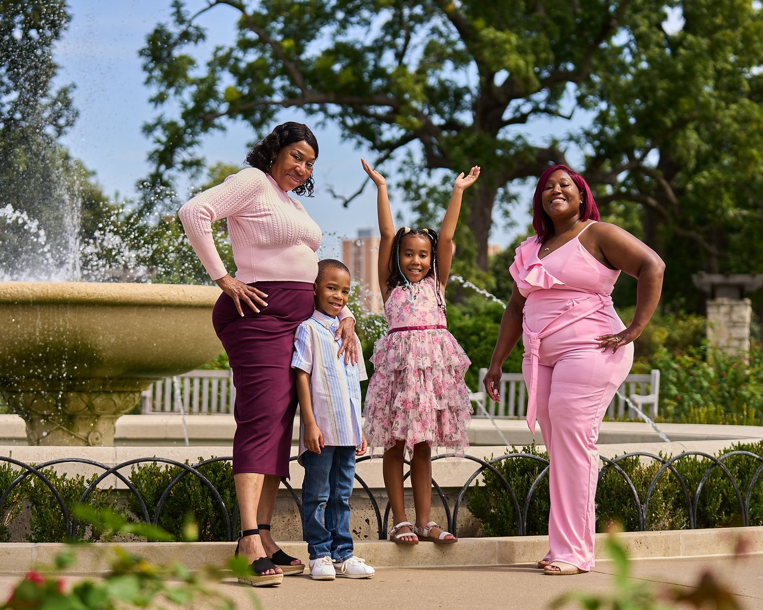 Family of four posing by a fountain in a park, wearing pink and purple outfits on a sunny day.