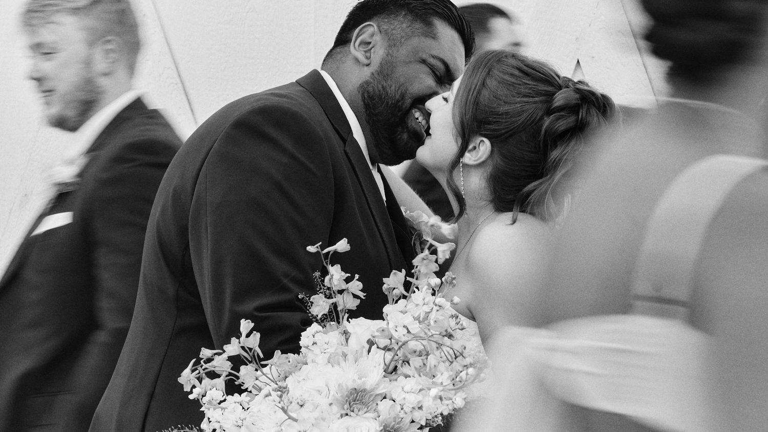 Black and white photo of a smiling groom in a suit leaning in to kiss his bride, who holds a bouquet of white flowers, as guests look on.