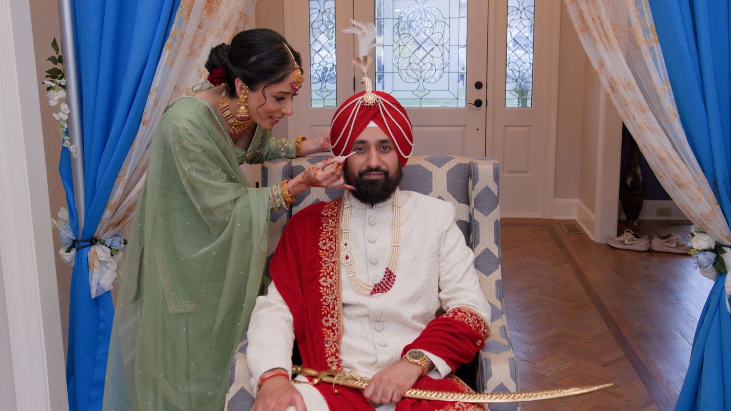 A woman in a green outfit adjusts the red turban of a seated groom in a cream and red sherwani holding a sword at a Sikh wedding.