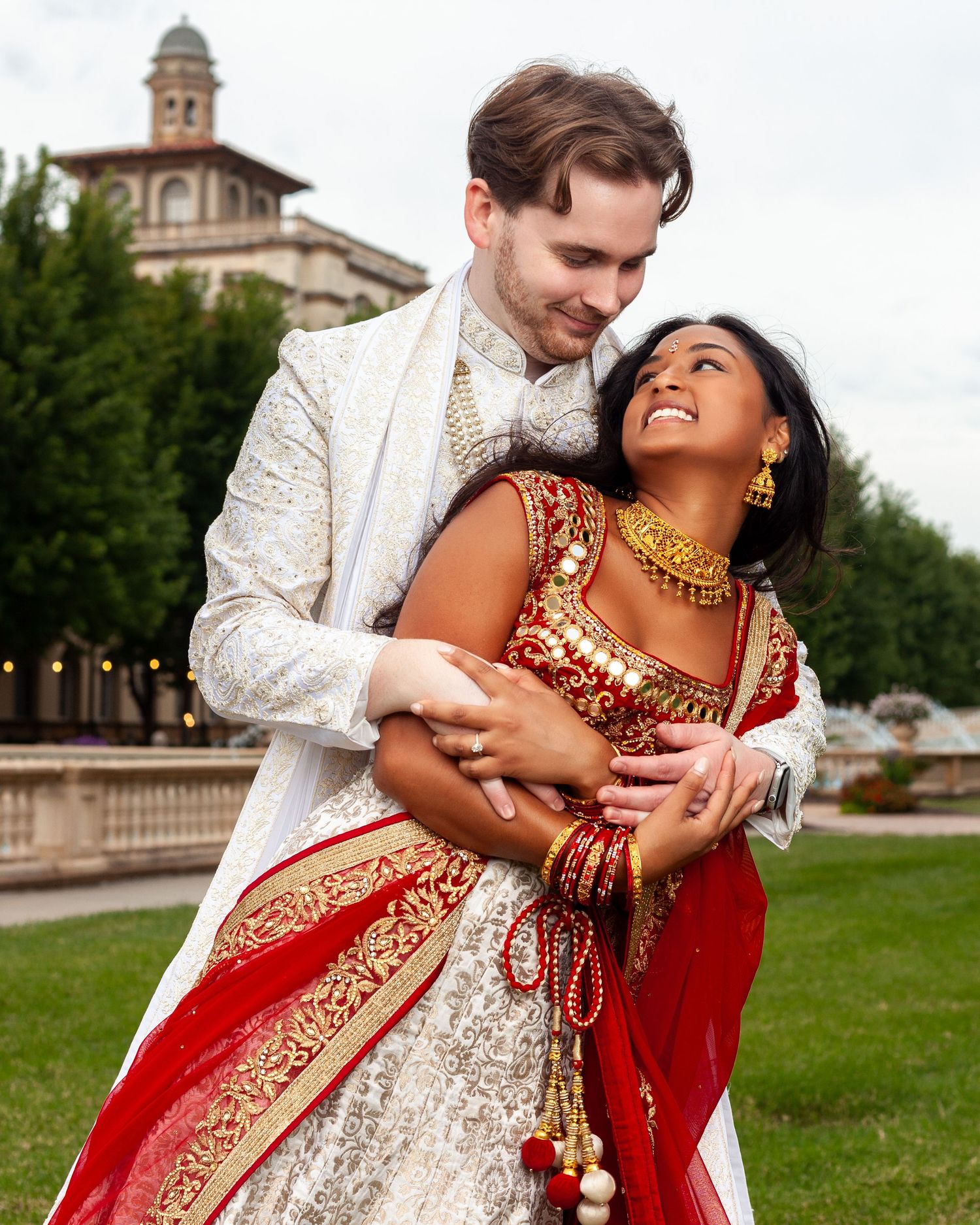 Couple in traditional Indian wedding attire embracing outdoors, bride in red and gold lehenga with jewelry.