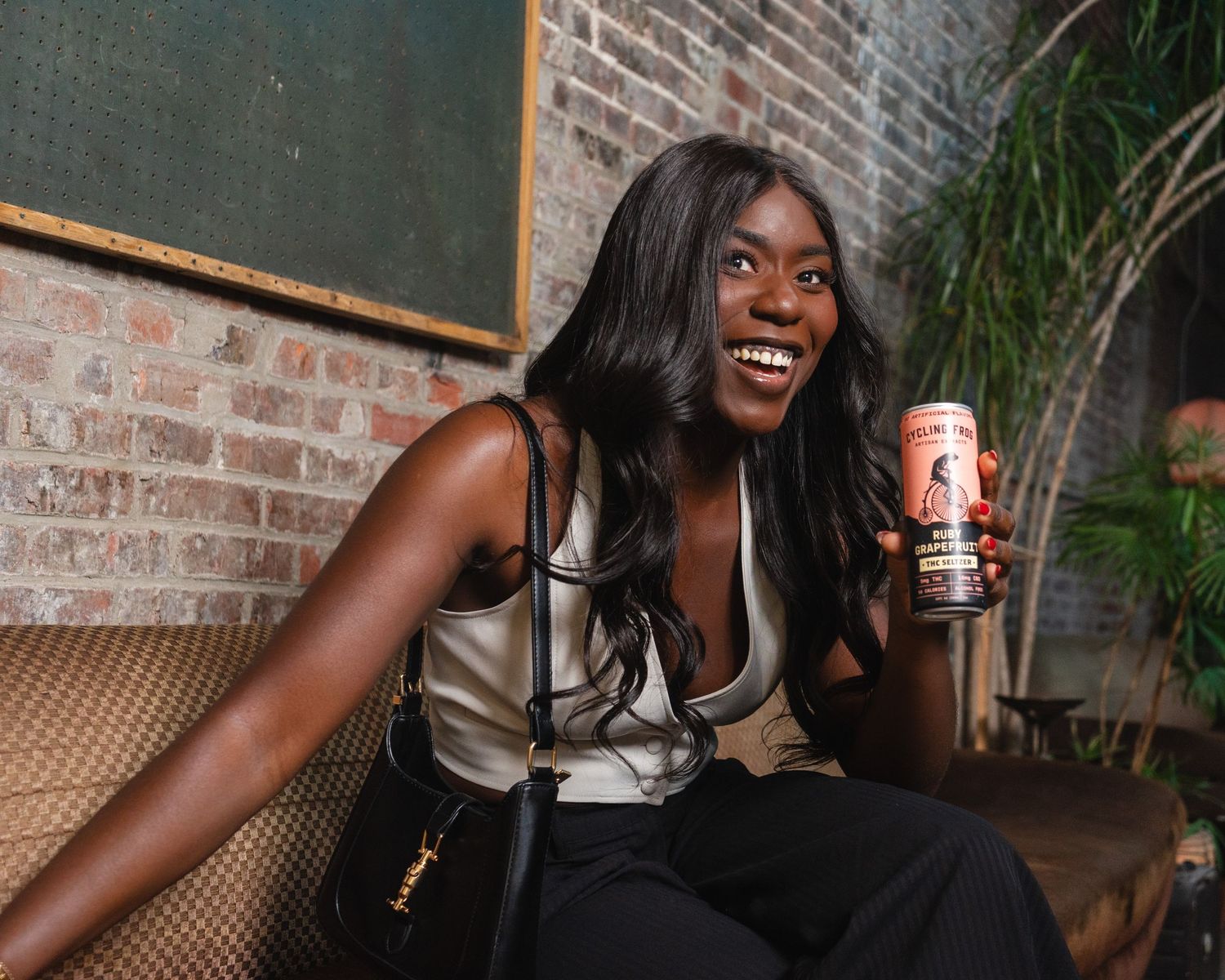 Smiling woman holding a canned beverage while seated in a stylish bar with brick walls and green decor.