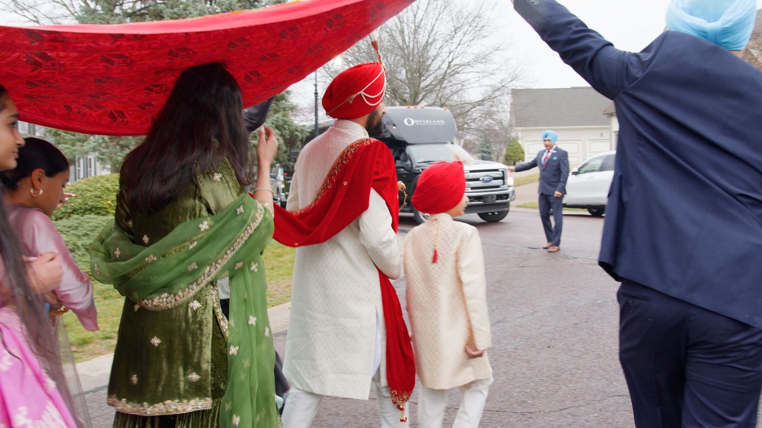 Sikh groom in ivory sherwani and red turban walks under a red canopy with a young boy and family during a wedding procession.