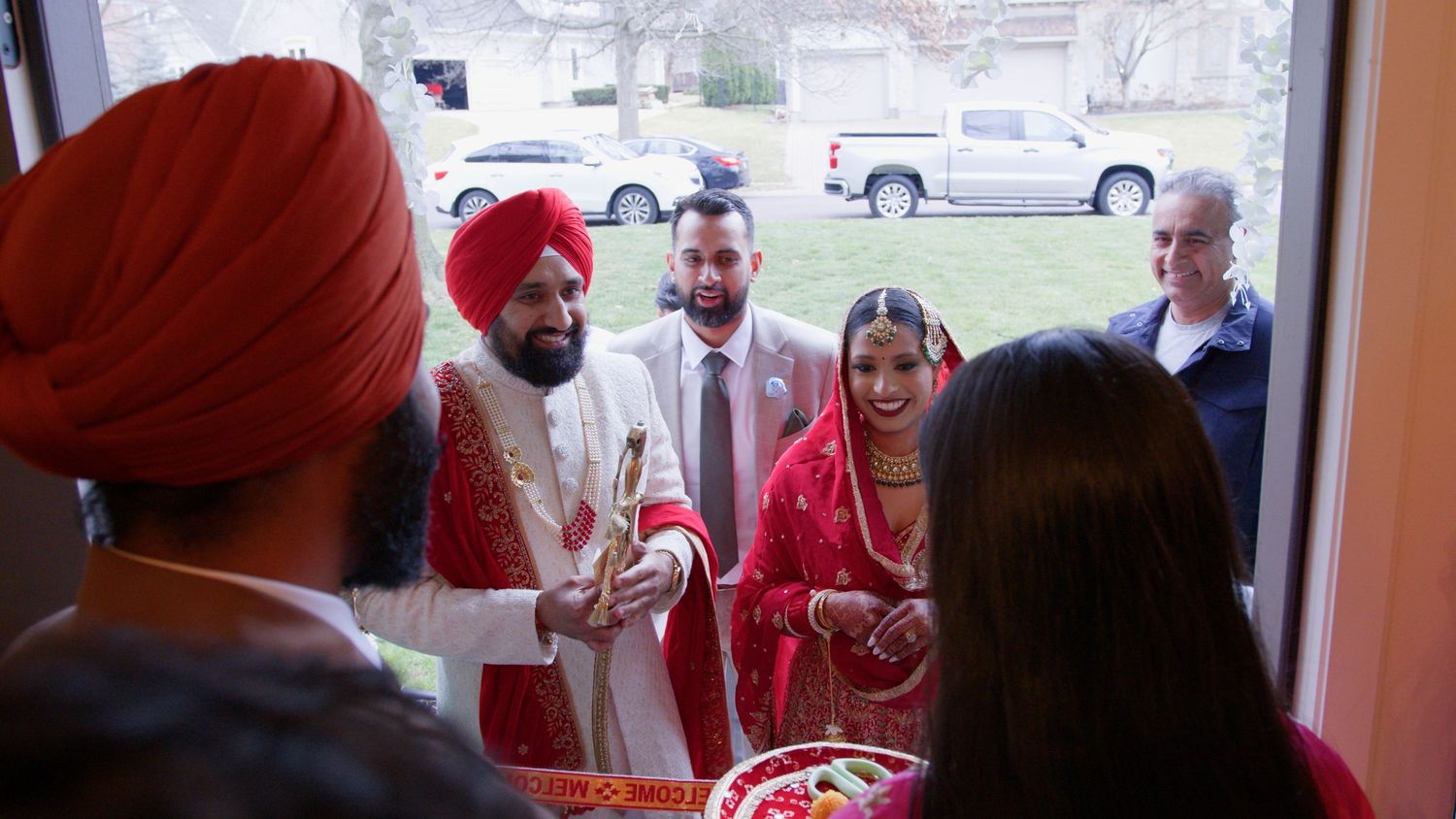 Groom in red turban and white sherwani with bride in red dress stand at a doorway during a traditional Punjabi wedding welcome ceremony.