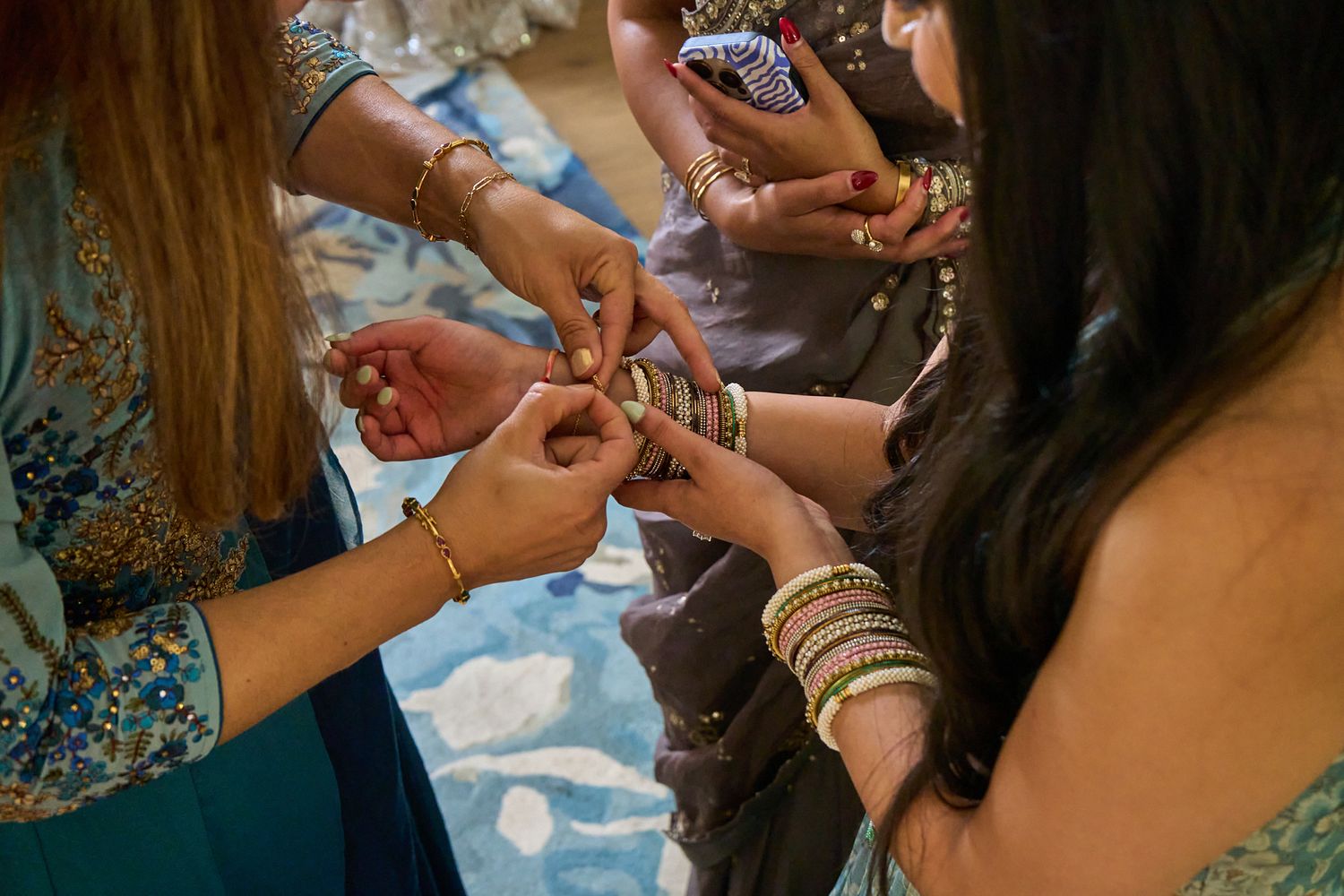 Two women in colorful embroidered outfits exchange and fasten gold and glass bangles on each other's wrists during a traditional ceremony.