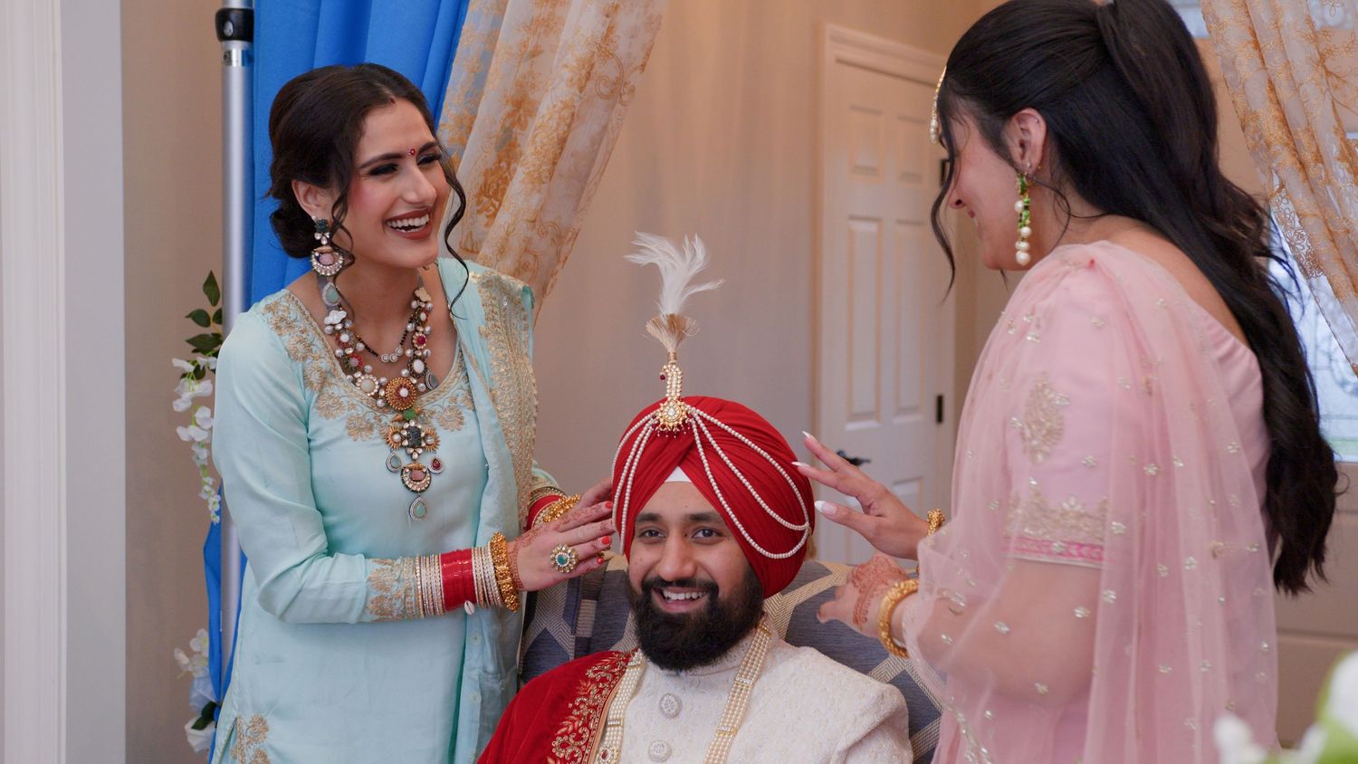 Two women in pastel Indian outfits adjust the red turban and feathered brooch of a smiling groom during a traditional Sikh wedding.