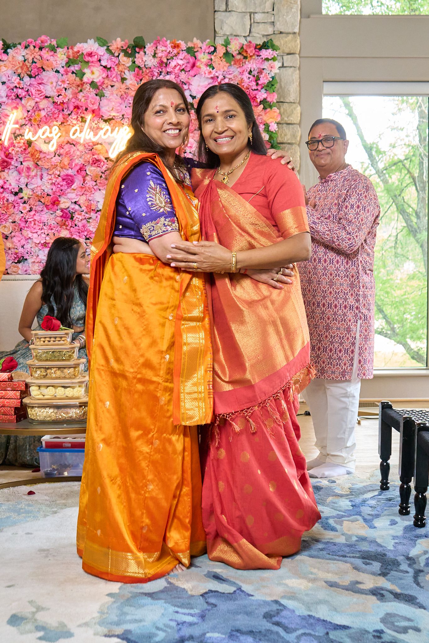 Two smiling women in orange and coral silk sarees embrace in front of a pink floral wall at a festive celebration.