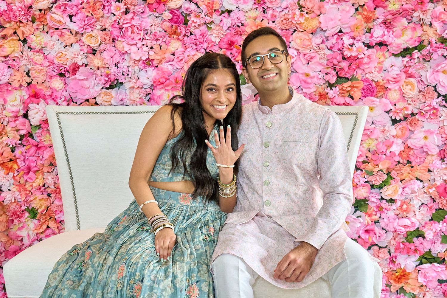 Smiling couple in traditional Indian attire sits on a white bench before a pink and orange floral wall, with the woman showing her ring.