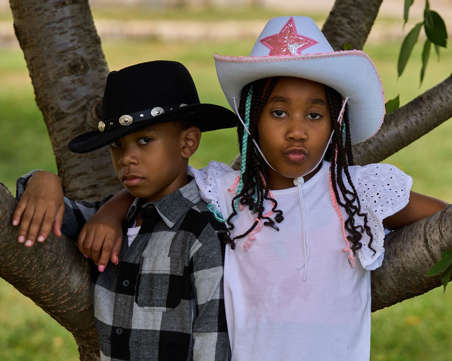 Two children wearing cowboy hats pose by a tree, boy in black hat and girl in pink star hat.