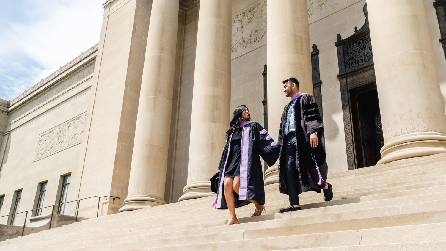 Two graduates in caps and gowns walk down steps of a neoclassical building, celebrating their academic achievement.