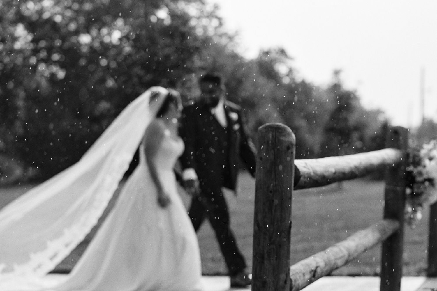 Black and white photo of a bride in a long veil and gown walking hand in hand with her groom beside a rustic wooden fence.