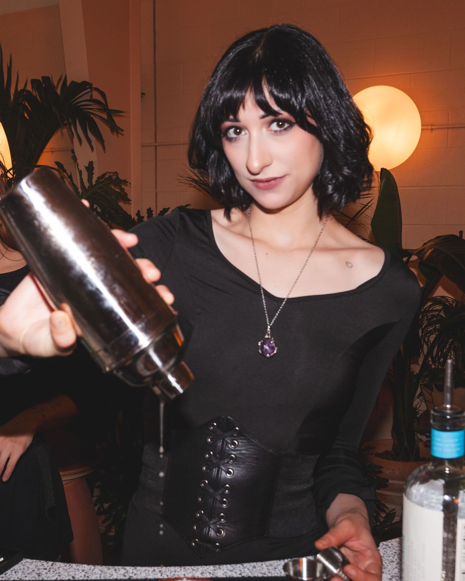 A bartender in black outfit pours a cocktail shaker at an indoor bar with warm ambient lighting.