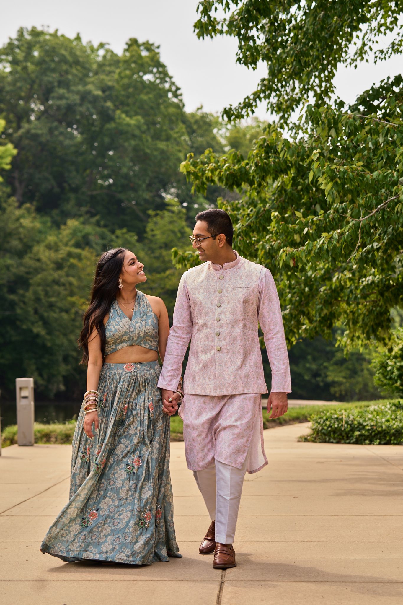 Indian couple in traditional attire walking hand in hand through a lush green park, smiling at each other.