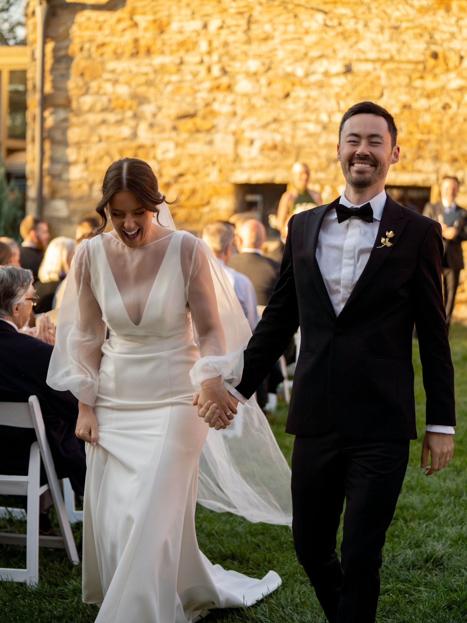 Joyful newlywed couple holding hands walking down outdoor aisle at rustic stone venue at sunset.