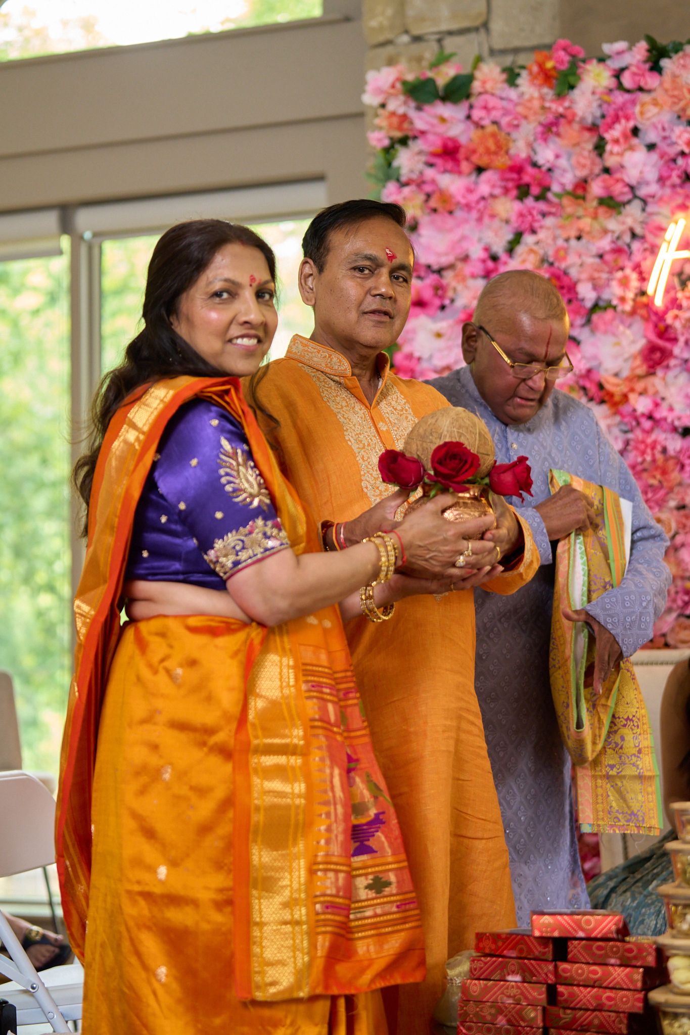 Smiling couple in traditional orange Indian attire holding red flowers at a ceremony, with a floral backdrop and an older man beside them.