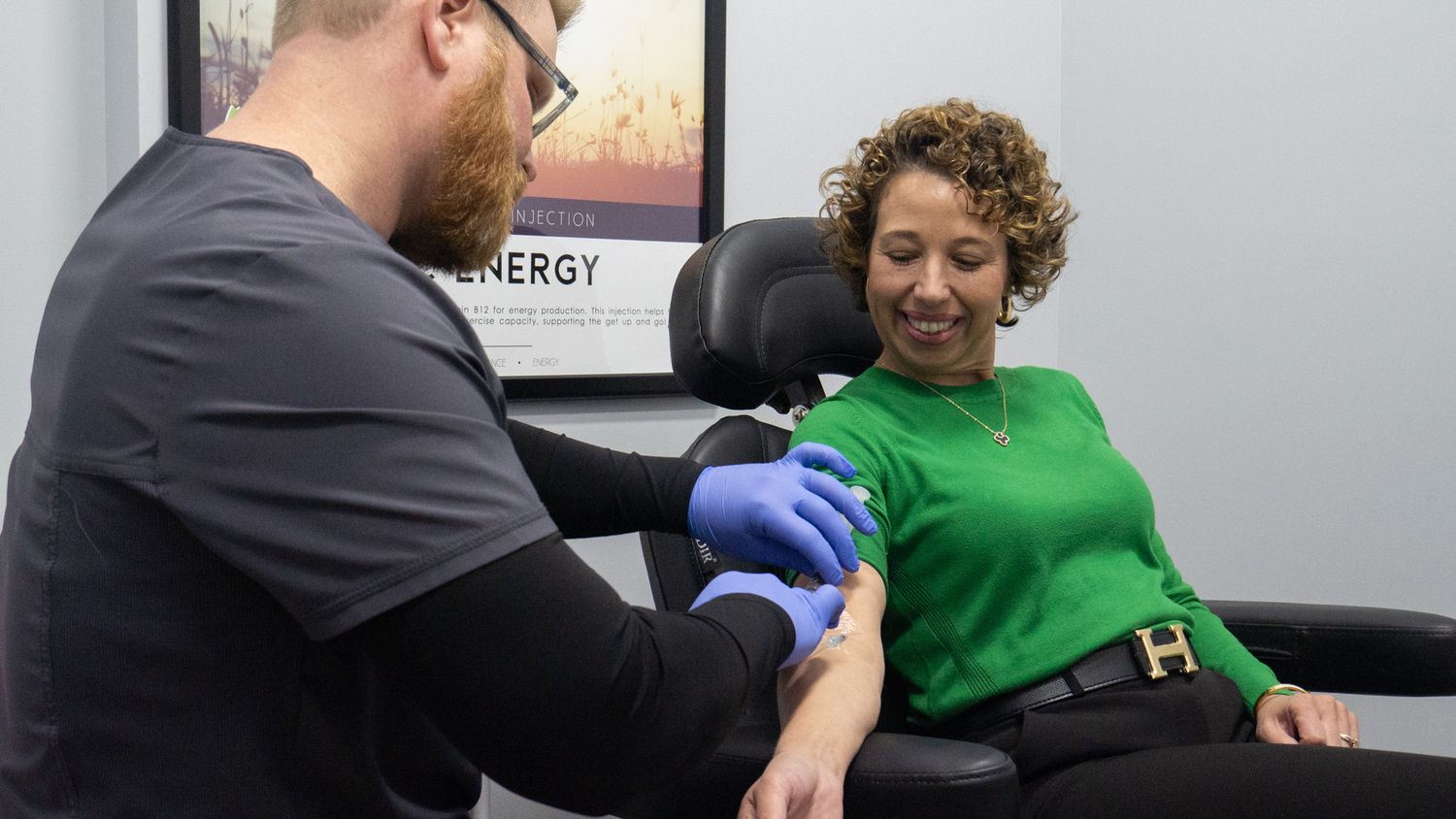 Healthcare professional in blue gloves performing a blood draw on a smiling woman in a green sweater at a medical clinic.