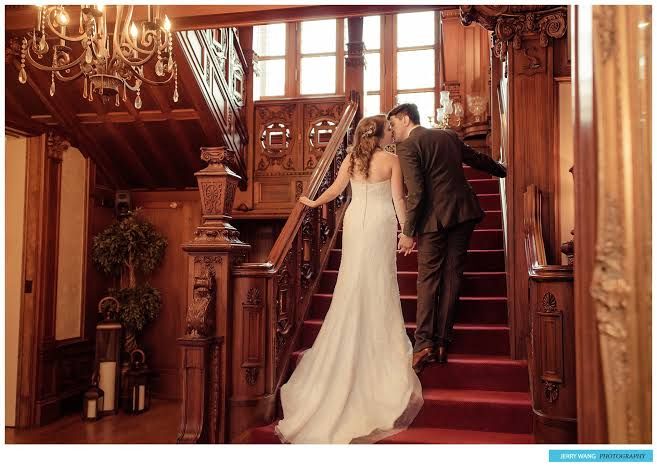 Bride in a flowing white gown and groom in a dark suit embrace on a red-carpeted grand wooden staircase beneath a crystal chandelier.