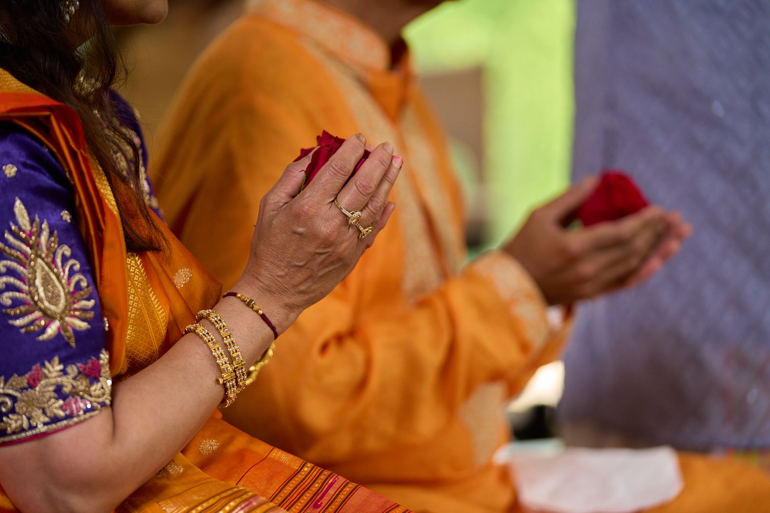 Two people in orange traditional Indian attire hold red flower petals in their hands during a ceremony, one wearing gold bangles and rings.
