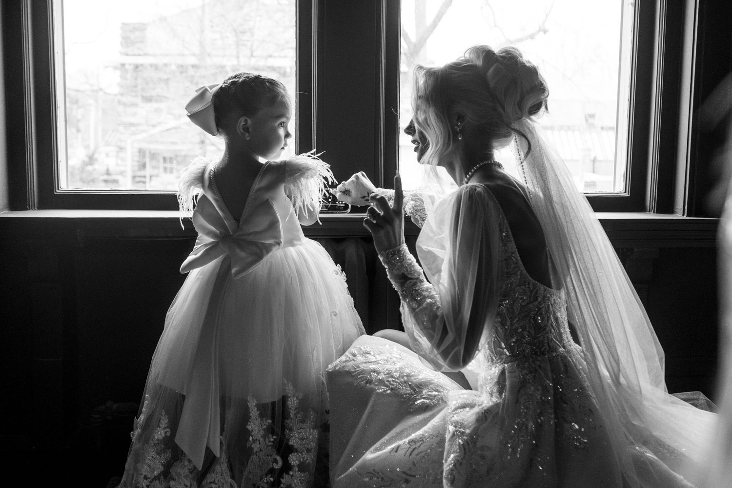 Black and white photo of a bride in lace gown sharing a tender moment with a flower girl by a window.