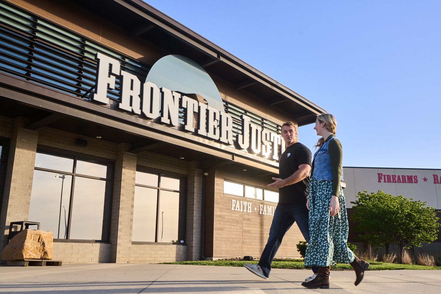Two people walking past Frontier Justice firearms store entrance on a sunny day.