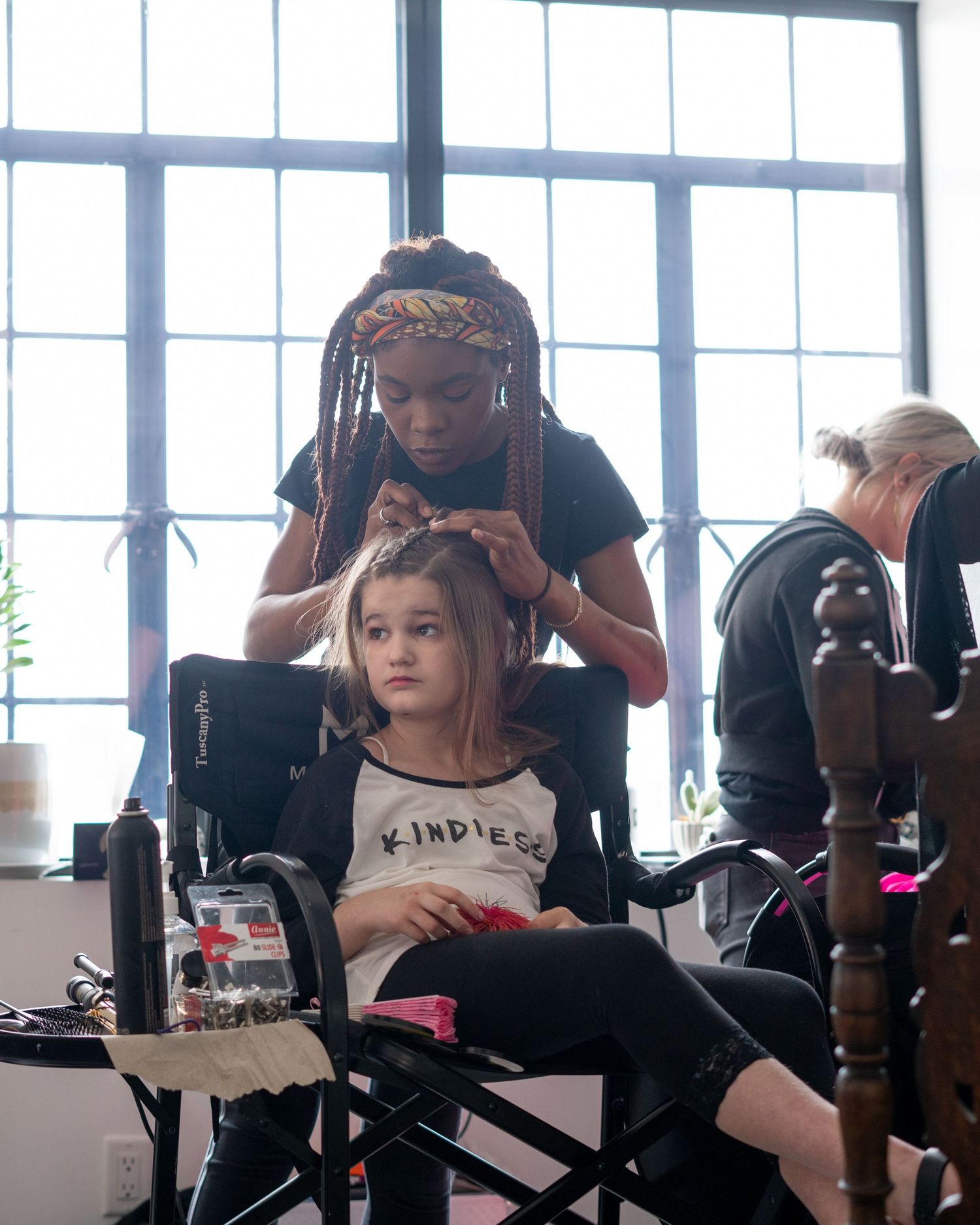 A hairstylist with dreadlocks styles a young girl's hair in a salon chair, girl wearing a Kindness shirt.