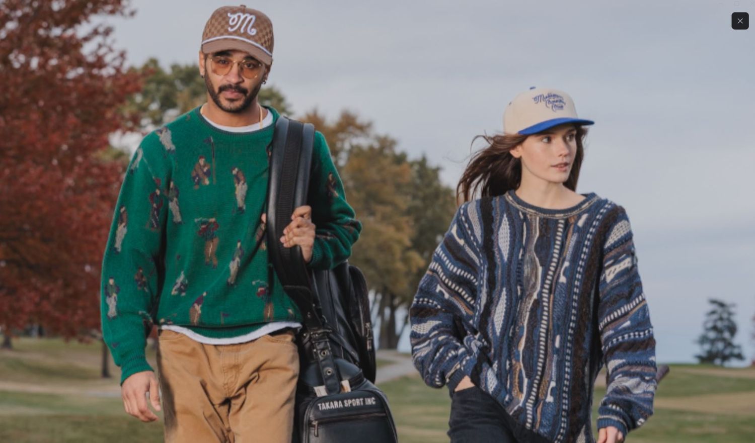 A man and woman walking outdoors in stylish casual fall outfits, wearing patterned sweaters and baseball caps.