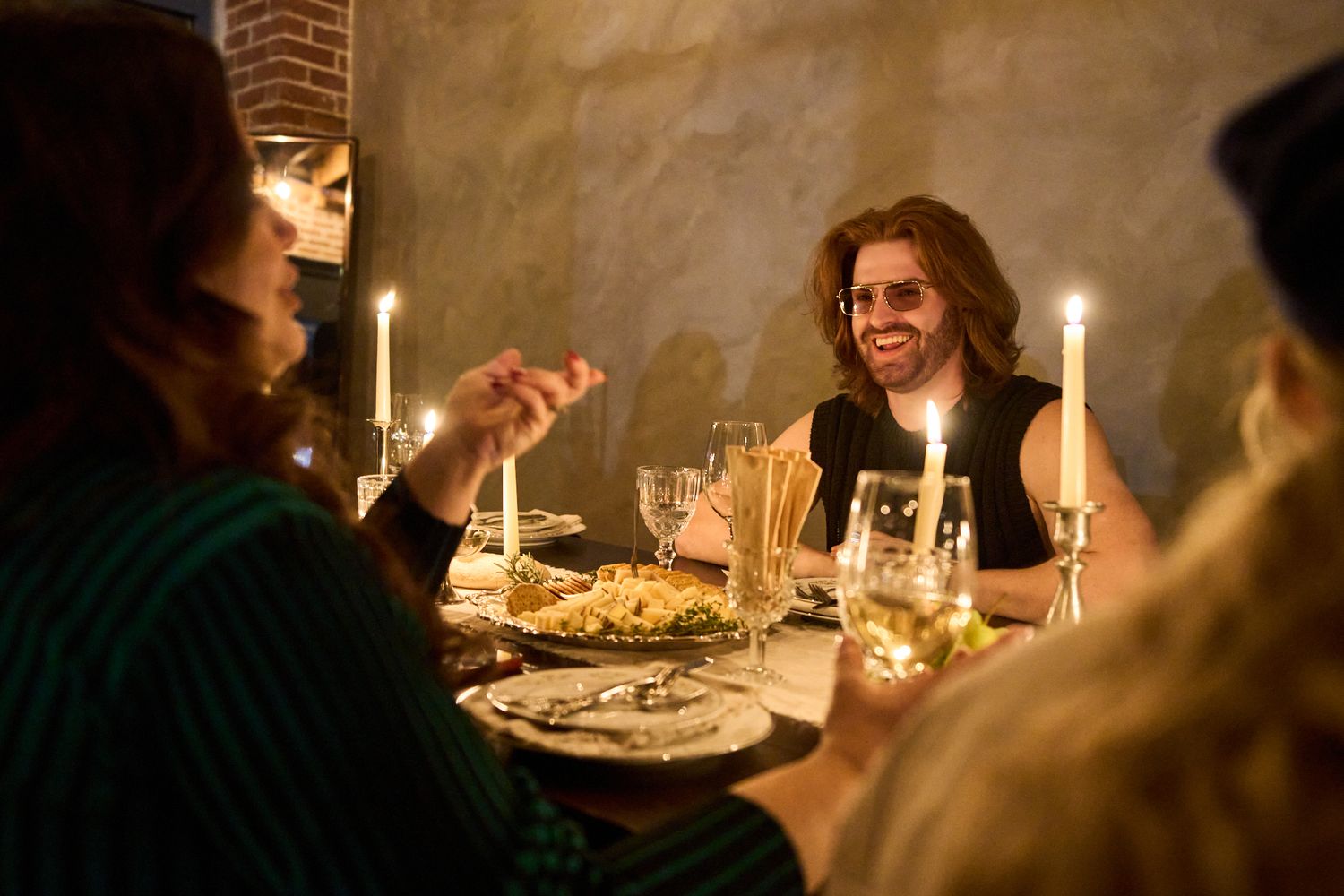 Friends enjoying a candlelit dinner party, smiling and conversing around a table with wine glasses and pasta dishes.