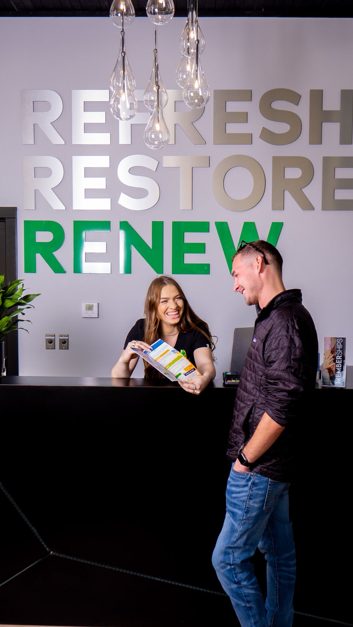 A friendly receptionist assists a male client at a modern wellness center desk with Refresh Restore Renew signage.