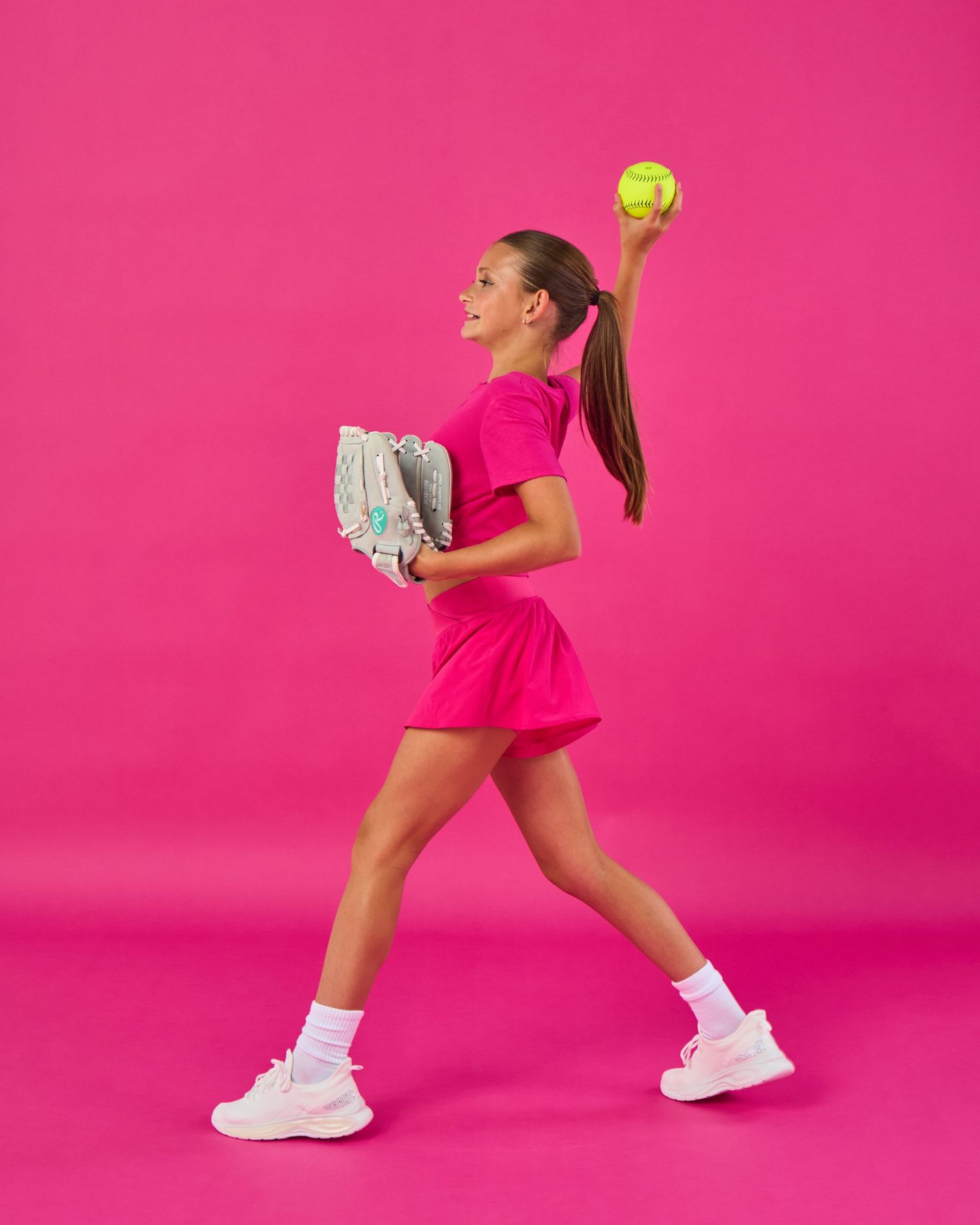 Young woman in pink dress holding a softball glove and raising a yellow ball against a pink background.