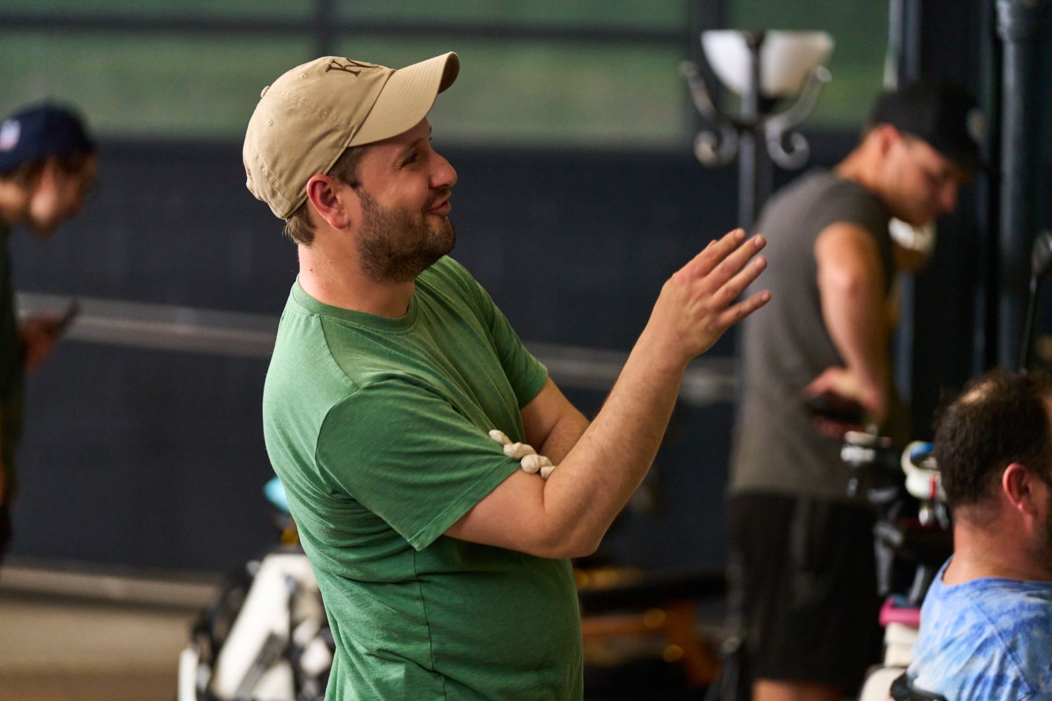 Man in green shirt and beige cap clapping hands on a film set with crew members in the background.