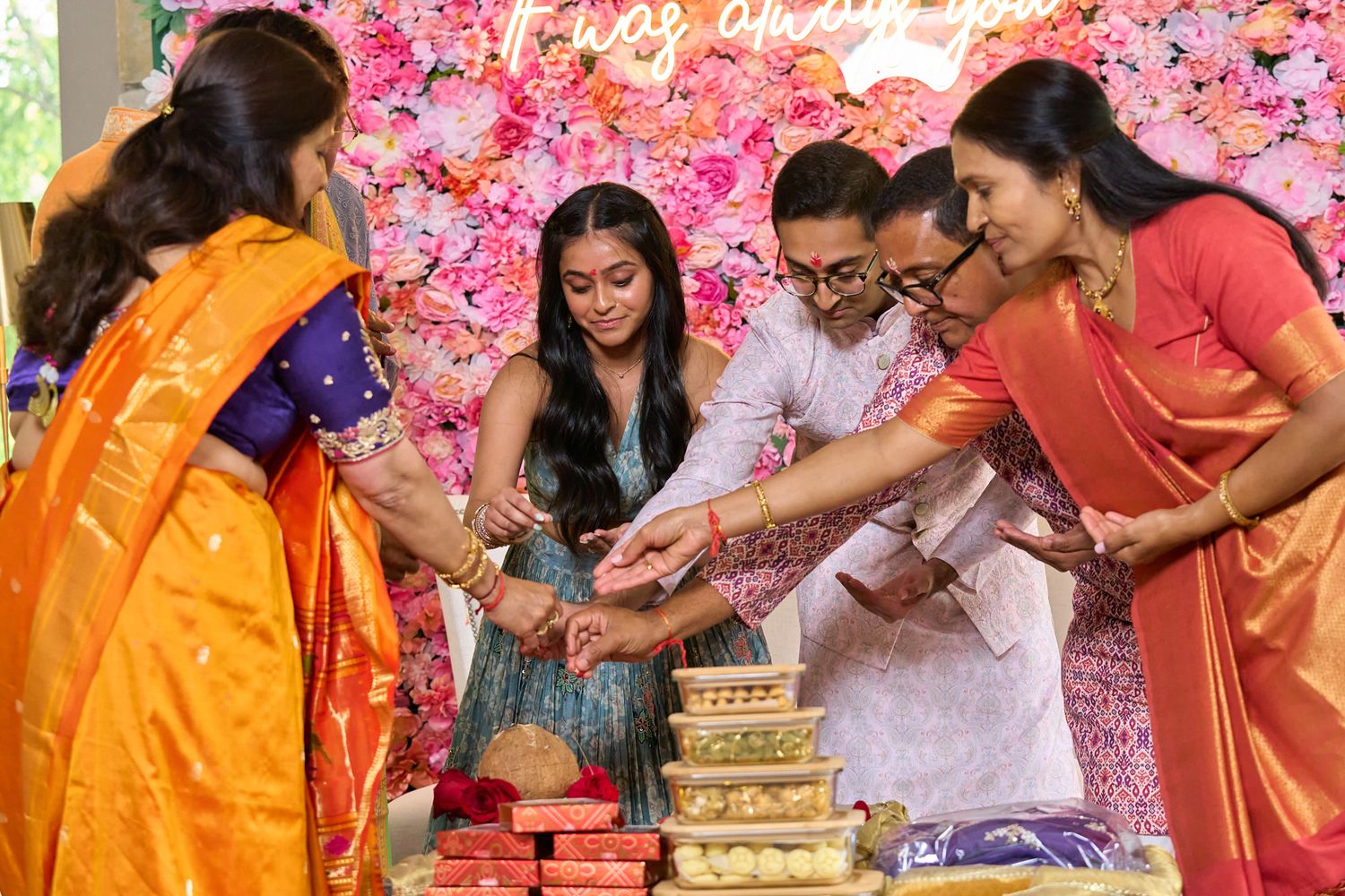 Indian family in colorful sarees sharing sweets at a festive celebration with floral backdrop.