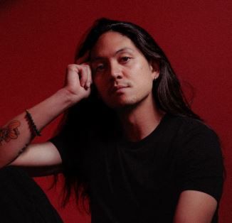 Long-haired person in a black t-shirt rests their head on a raised hand, with arm tattoos and a bracelet, against a bold red backdrop.