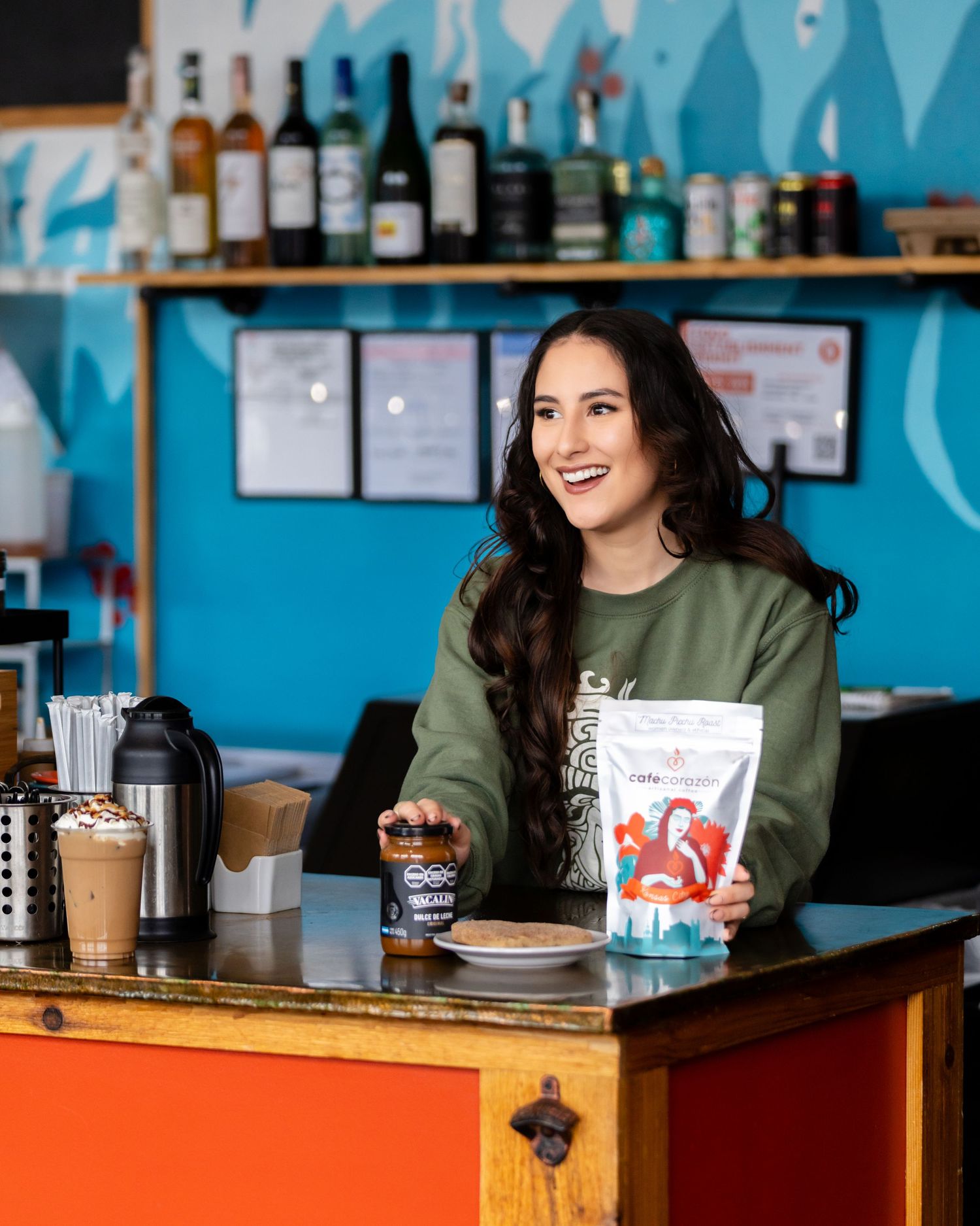 Woman smiling behind a cafe counter holding snack products, with bottles and a blue wall in the background.