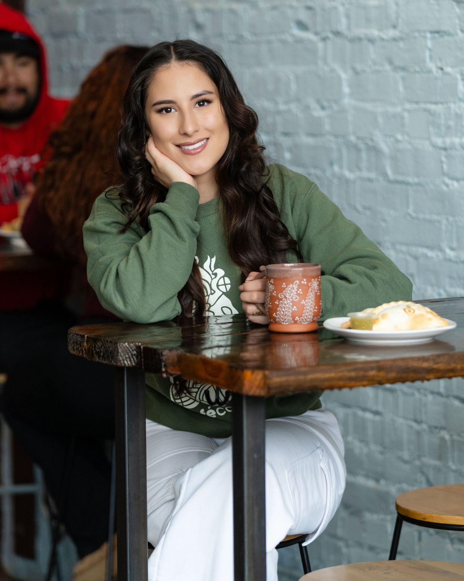 Smiling young woman in green sweatshirt sitting at wooden cafe table with clay mug and plate of food.