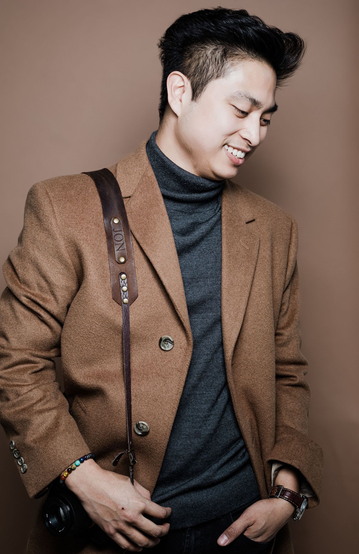 Stylish Asian man in brown blazer and gray turtleneck smiling with camera strap over shoulder.