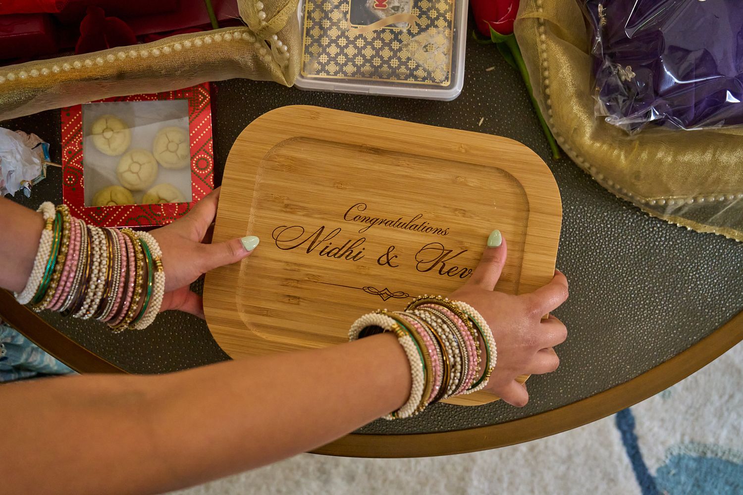 Hands adorned with colorful bangles hold a wooden engraved tray reading Congratulations Nidhi & Ravi at a traditional wedding ceremony.