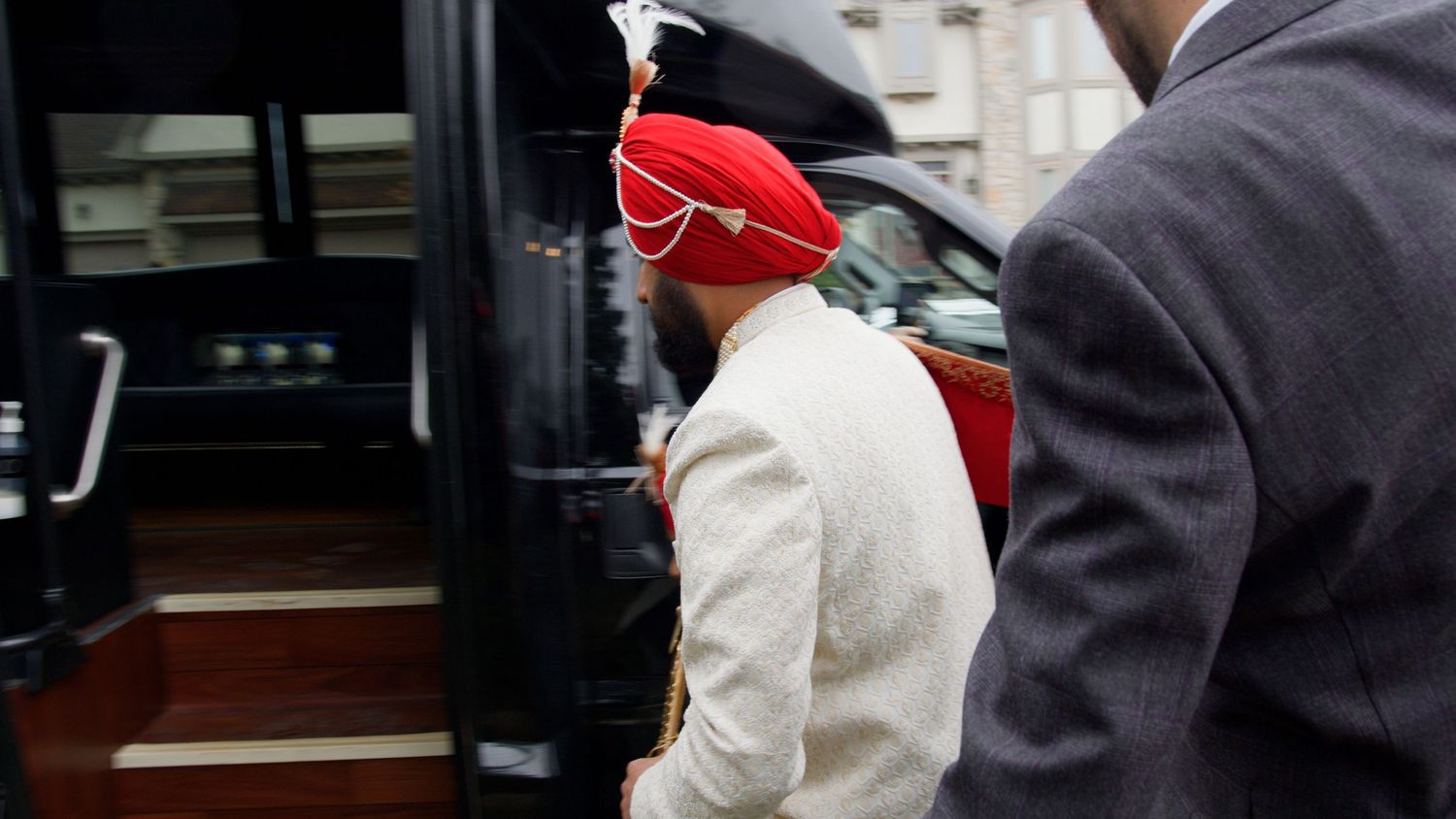 Man in a red turban and cream jacket with a pearl sash steps into a black vehicle while another man in a gray suit stands nearby.